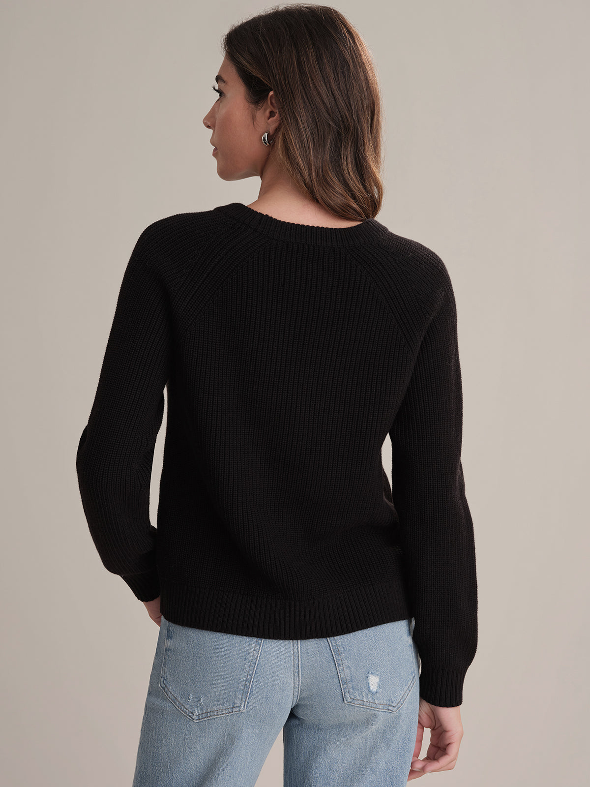 A person stands facing away from the camera, wearing a 525 America black cotton raglan-sleeve sweater and light blue jeans with a small rip on the back pocket against a plain, neutral background.