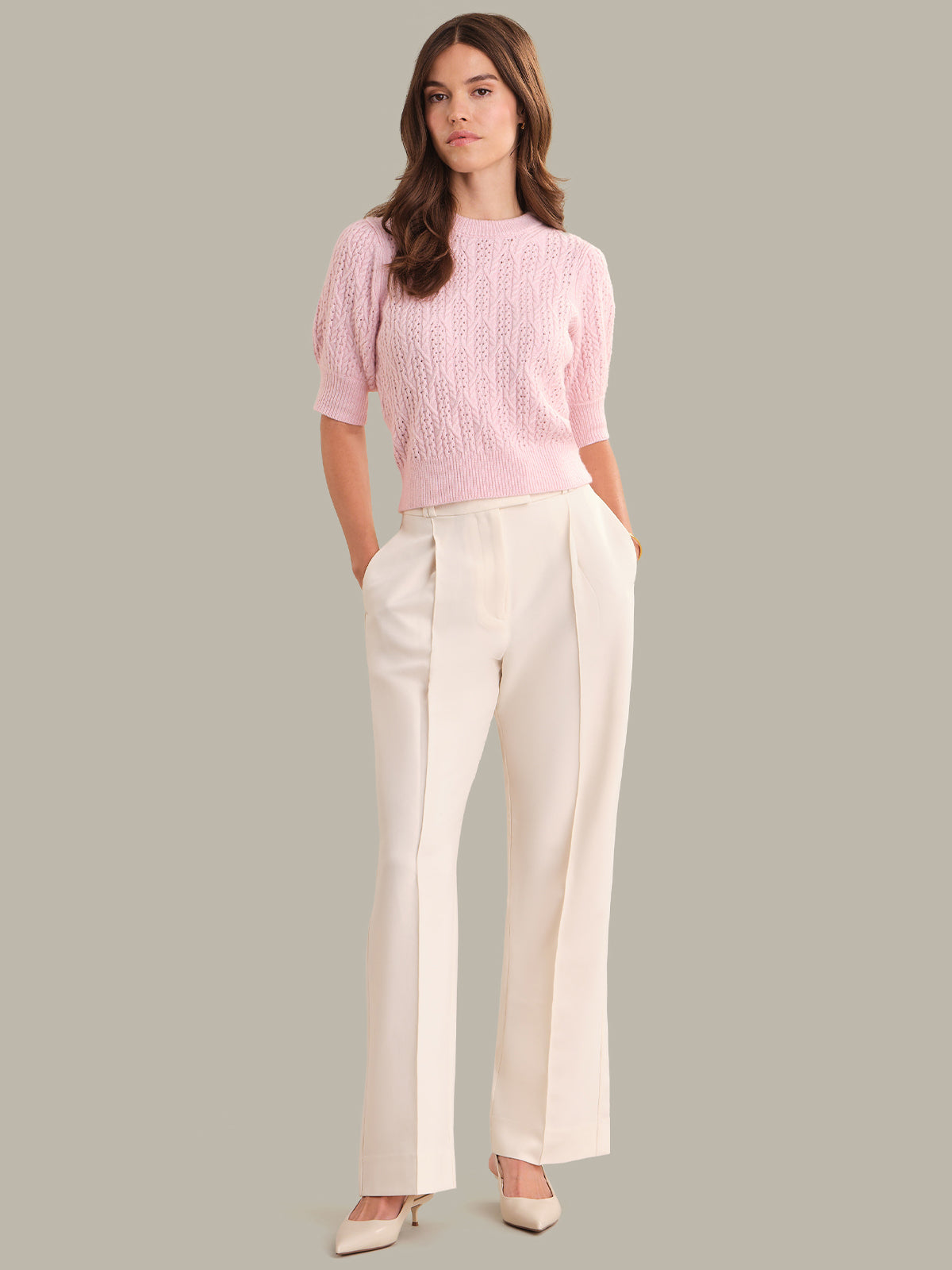 Woman wearing a pink knitted top and white pants against a gray background
