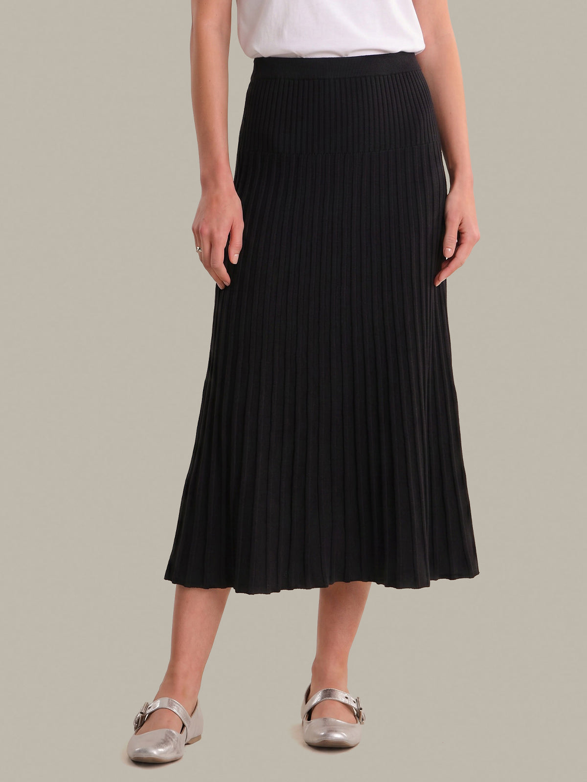 A model wearing a black pleated midi skirt, styled with a white top and metallic silver flats, set against a solid light gray background.