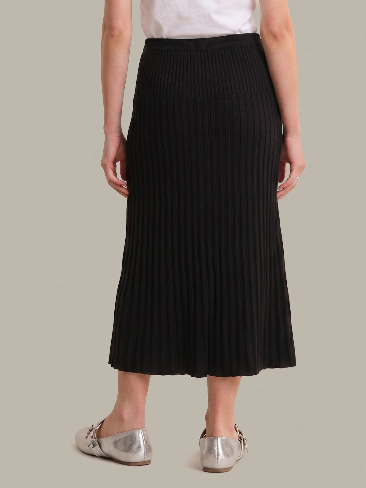 A model from the back wearing a black pleated midi skirt, a white top, and metallic silver flats, set against a solid light gray background.