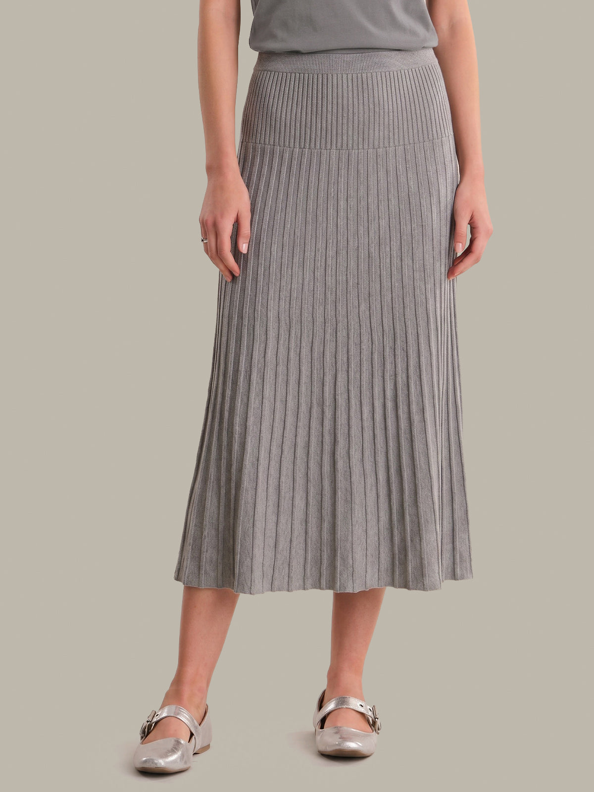 A model wearing a gray pleated knit midi skirt with a ribbed waistband, paired with a matching gray top and metallic silver flat shoes, set against a solid light gray background.