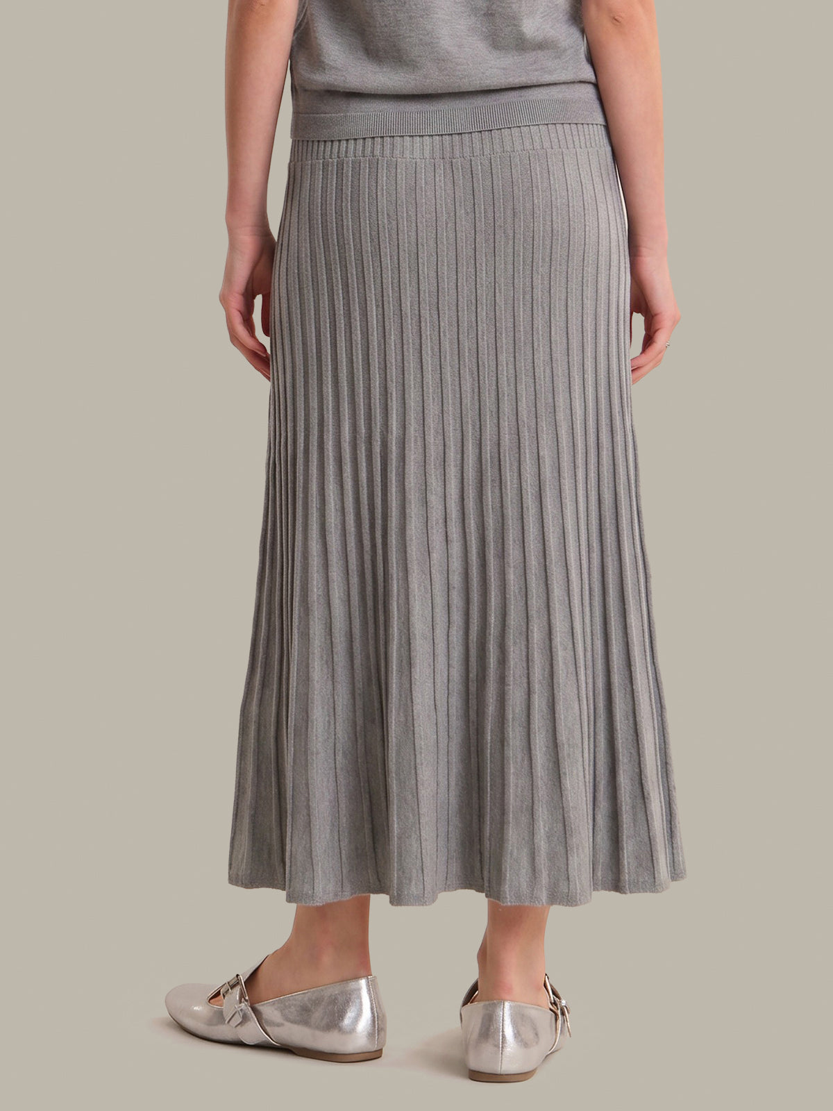 A model from the back wearing a gray pleated knit midi skirt, a matching gray top, and metallic silver flats, set against a solid warm gray background.