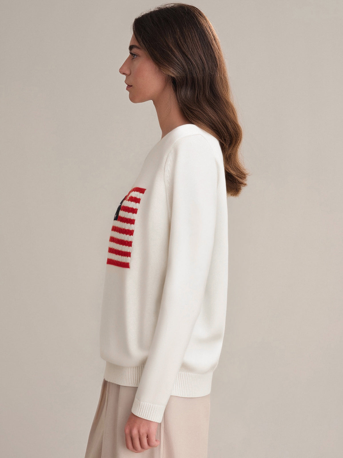 A woman with long brown hair stands in profile against a plain background, wearing a 525 America 100% cotton crewneck white sweater with a red and blue USA flag design on the front, paired with a beige skirt.