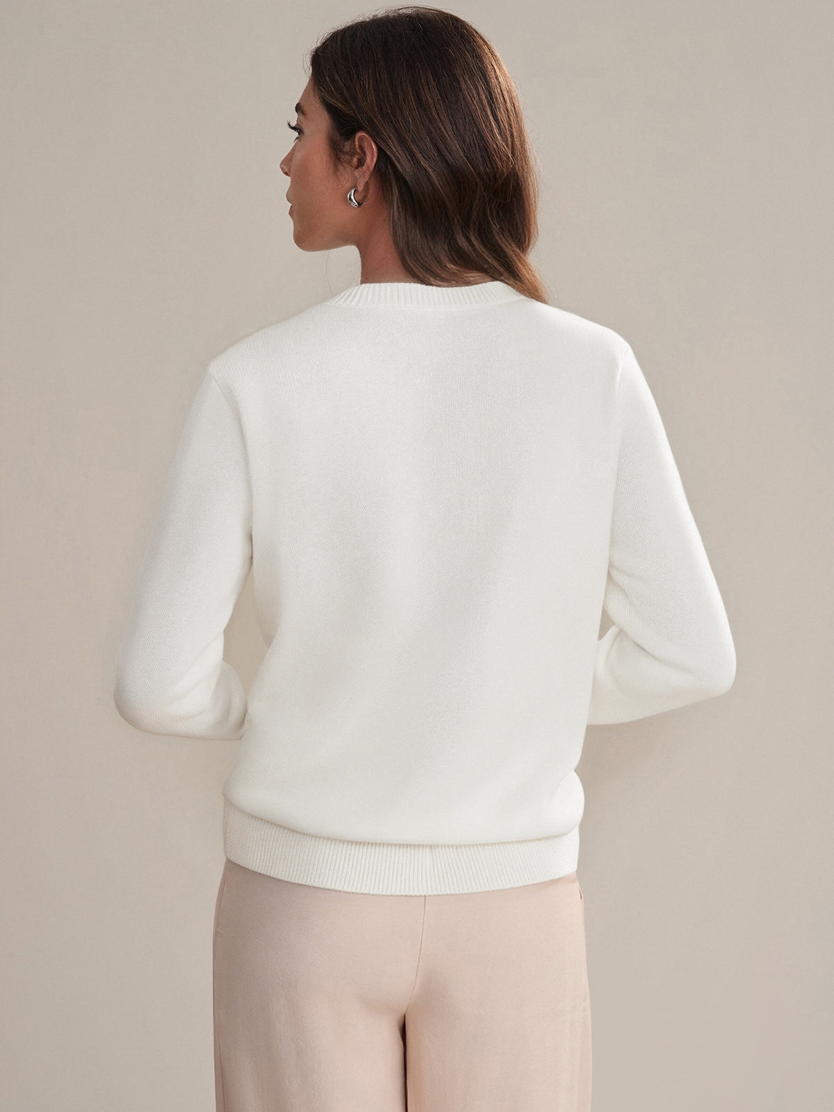A person with long brown hair is seen from behind, wearing a 525 America 100% cotton white crewneck sweater and light beige pants against a plain, neutral background.