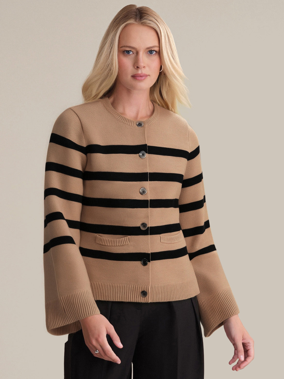 A woman with long blonde hair exudes sophistication in a timeless 525 America stripe cardigan with black horizontal stripes, round neckline, button closure, and two front pockets, paired with black pants against a plain light background.