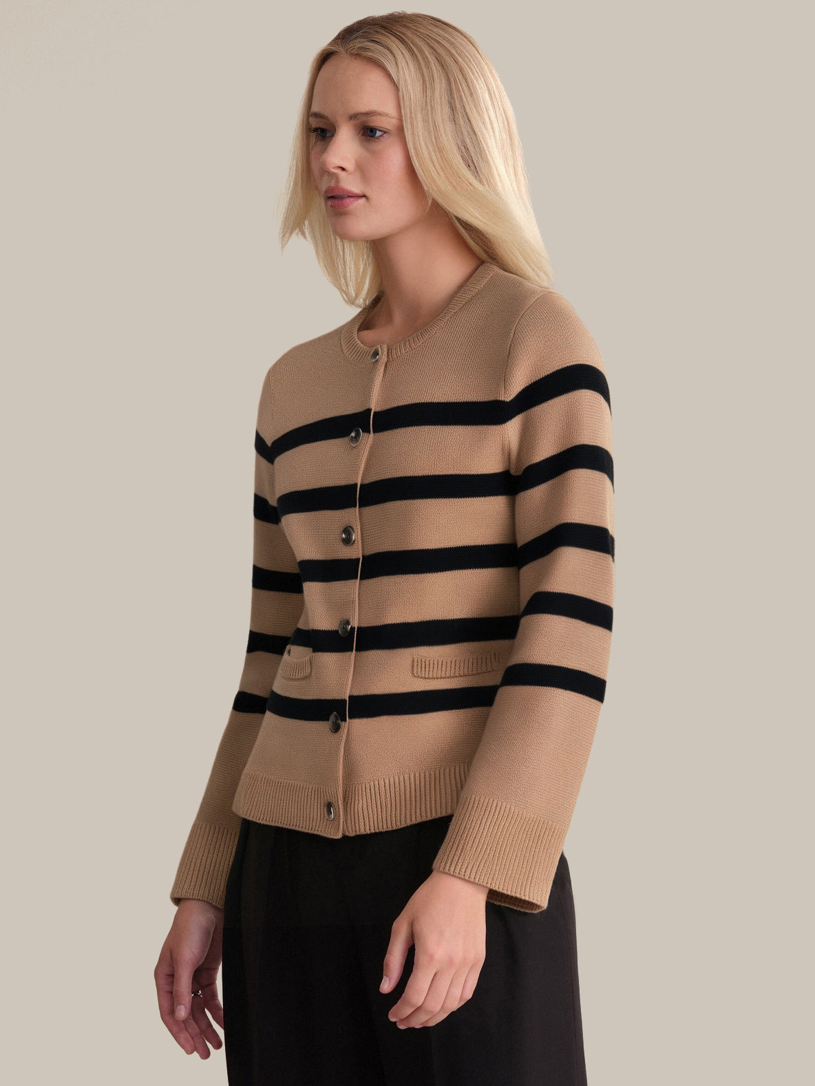 A person with long blond hair wears a 525 America timeless camel stripe cardigan with black buttons and black pants, standing against a plain beige background.