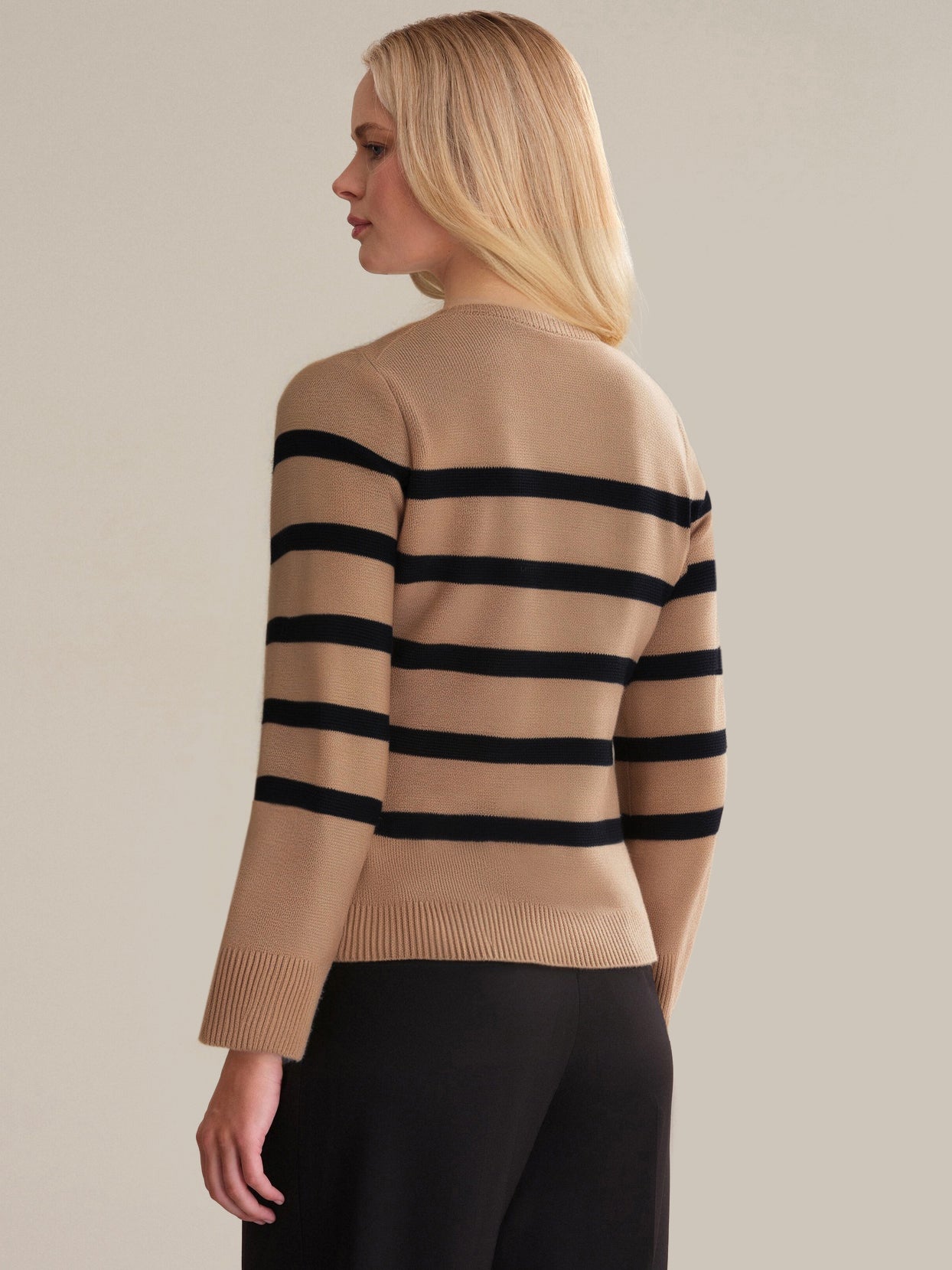 A woman with straight blonde hair stands facing away, exuding sophistication in a timeless 525 America stripe cardigan and black pants against a plain, neutral background.