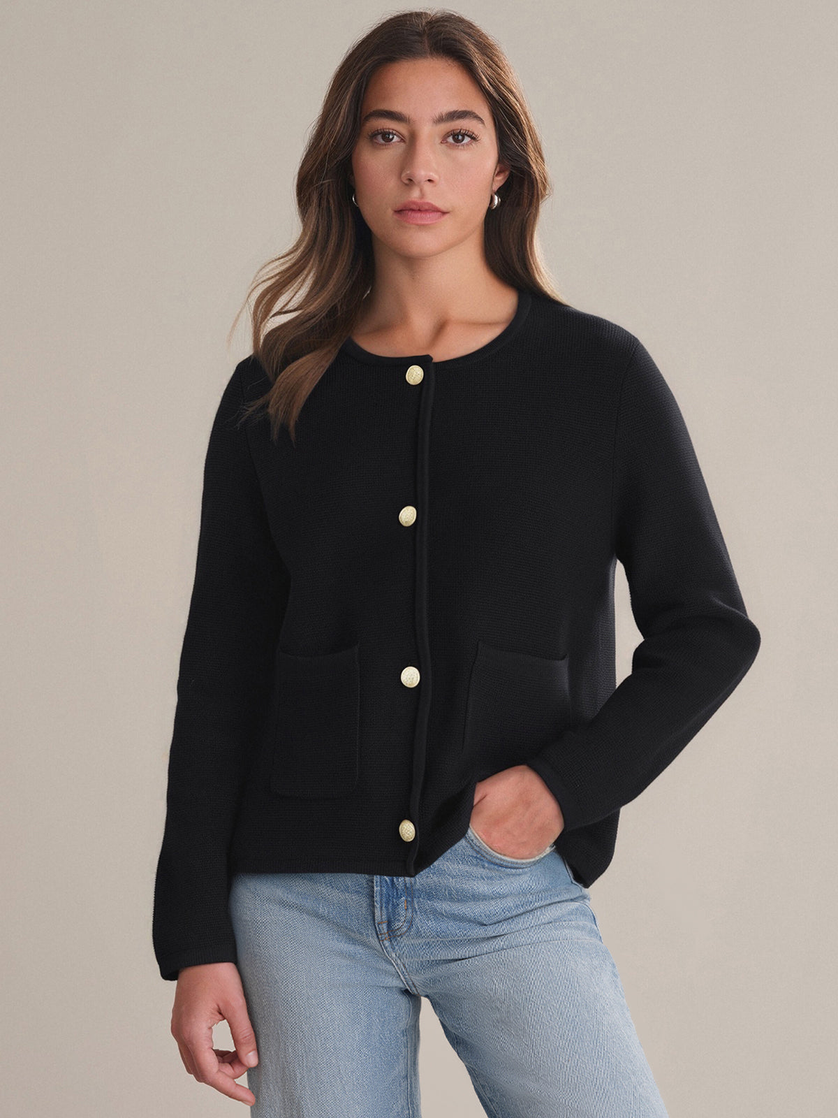 A person with long, wavy brown hair and a neutral expression wears the 525 America black pure cotton crewneck cardigan with gold buttons and front pockets, paired with light blue jeans. This piece is a timeless style addition to any wardrobe.