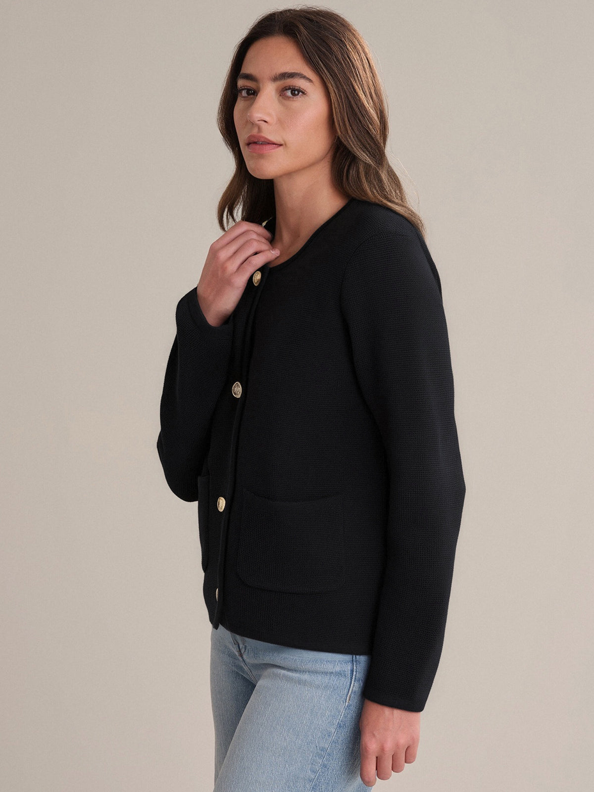 A person with long brown hair wears a 525 America black crewneck cotton cardigan with large front pockets and light blue jeans, standing against a plain light background—a versatile addition to any wardrobe.