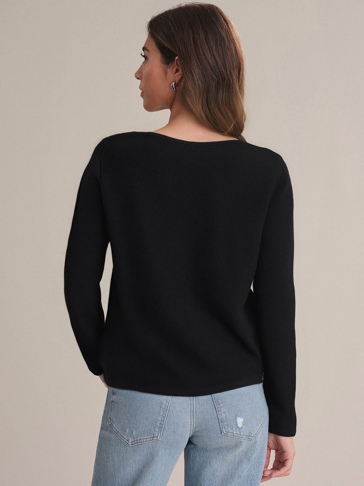 A person with long brown hair is seen from the back, wearing a 525 America pure cotton black crewneck cardigan and light blue jeans against a beige background—a timeless wardrobe essential.