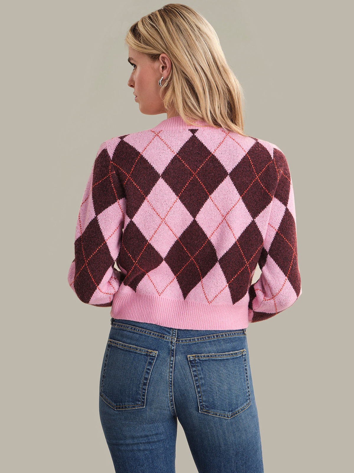 Person wearing a pink and brown argyle sweater with blue jeans on a plain background
