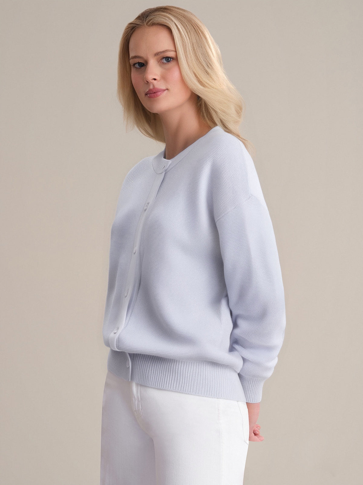 A woman with long blonde hair stands against a beige background, wearing a 525 America light blue knit button-up cardigan with a crew neckline and white pants. She looks slightly to the side with a neutral expression.