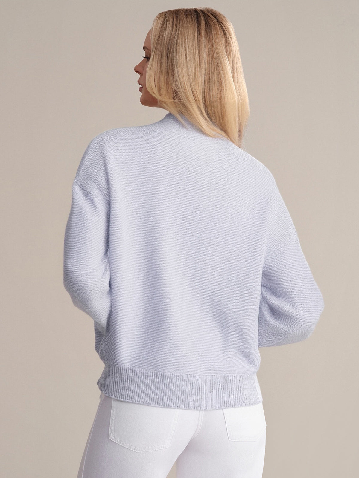 A person with long, straight blond hair is seen from the back, wearing a lightweight lavender crewneck sweater by 525 America and white pants against a plain neutral background.