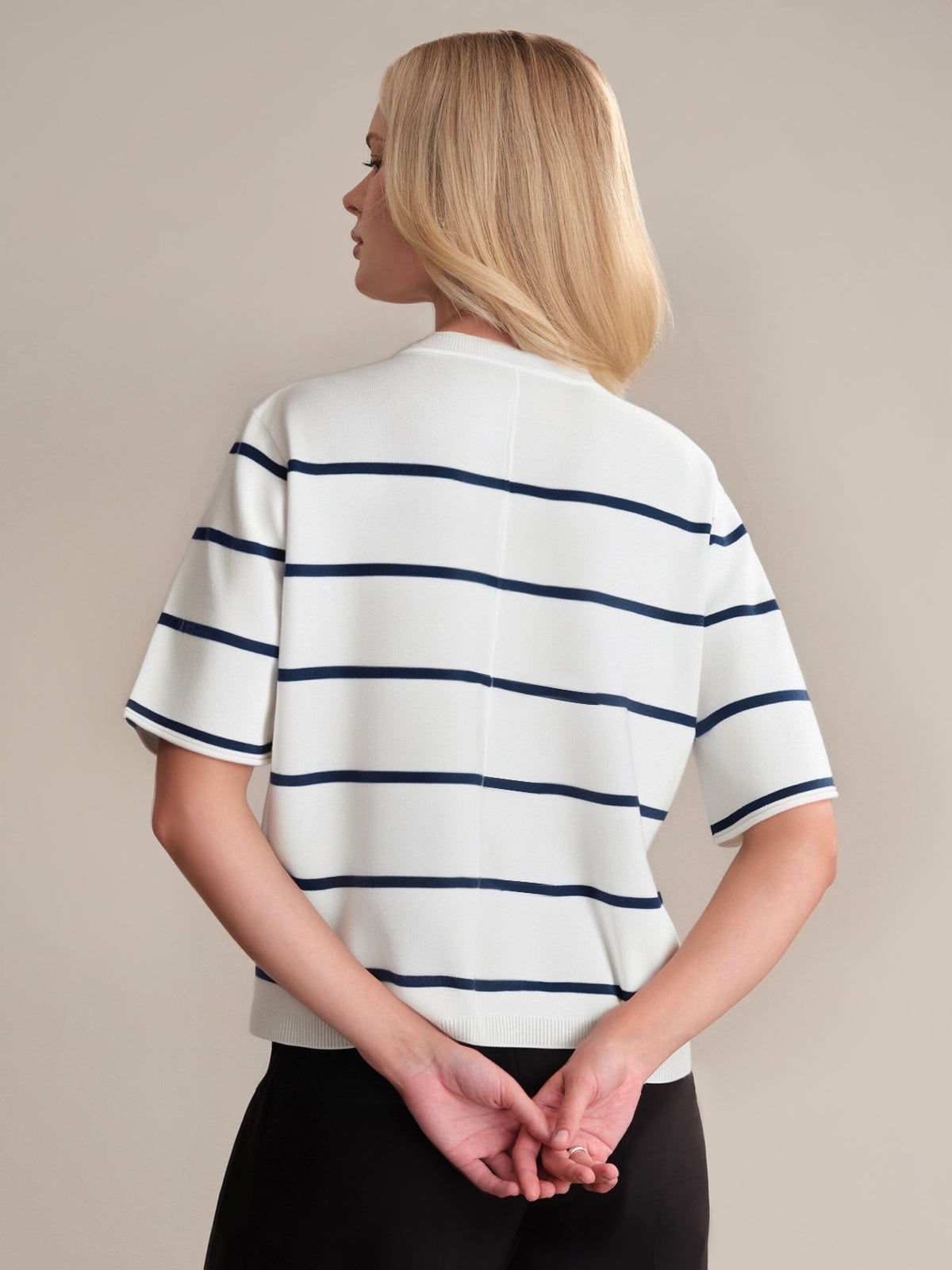 A person with blonde, shoulder-length hair stands facing away, wearing a 525 America premium cotton sweater with horizontal navy blue stripes and black pants, hands clasped behind their back against a plain background.