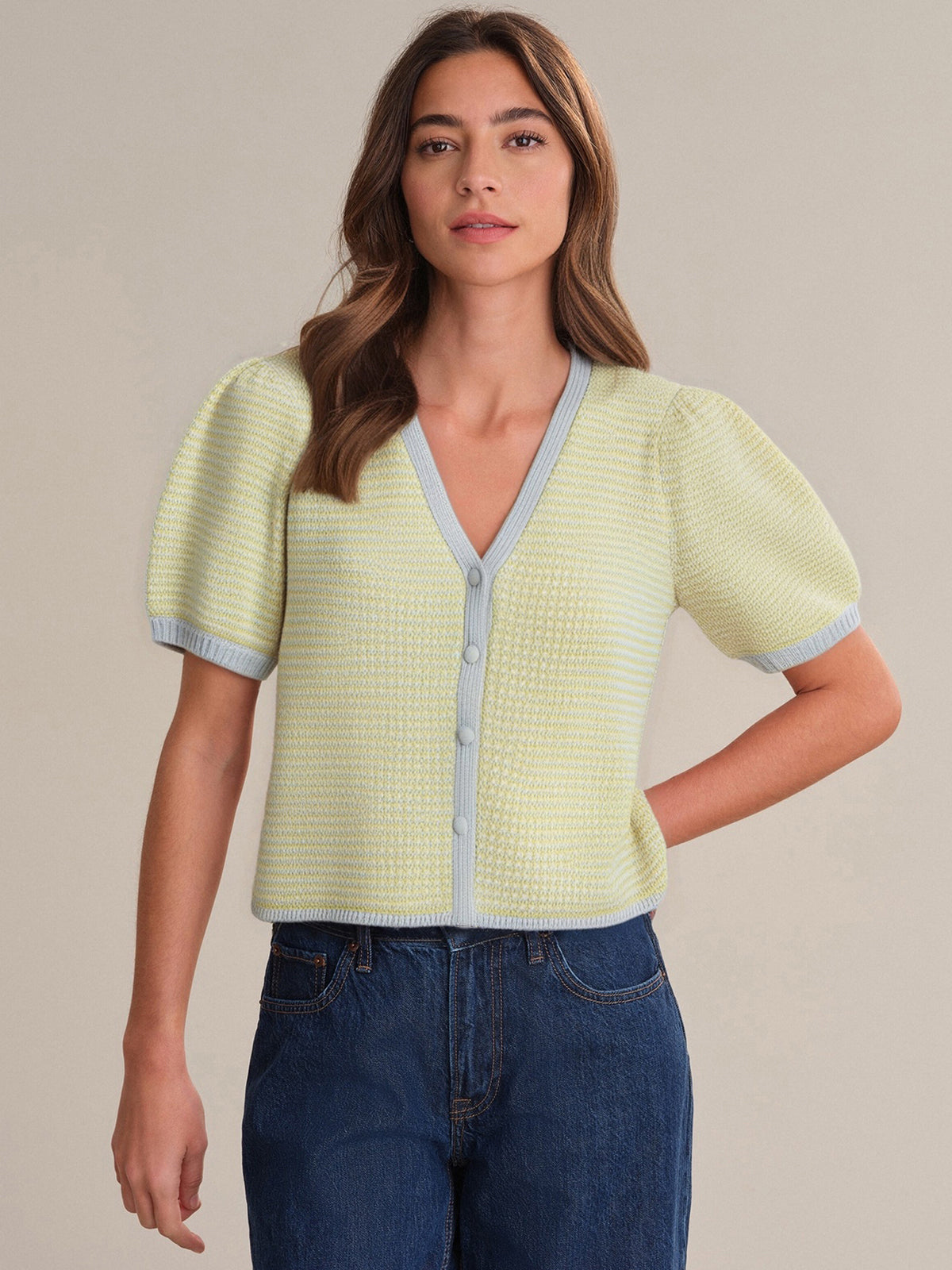 A woman with long brown hair wears a 525 America light yellow 100% cotton knit sweater featuring puff sleeves, light blue trim, and V-neck button front, paired with blue jeans against a plain background.