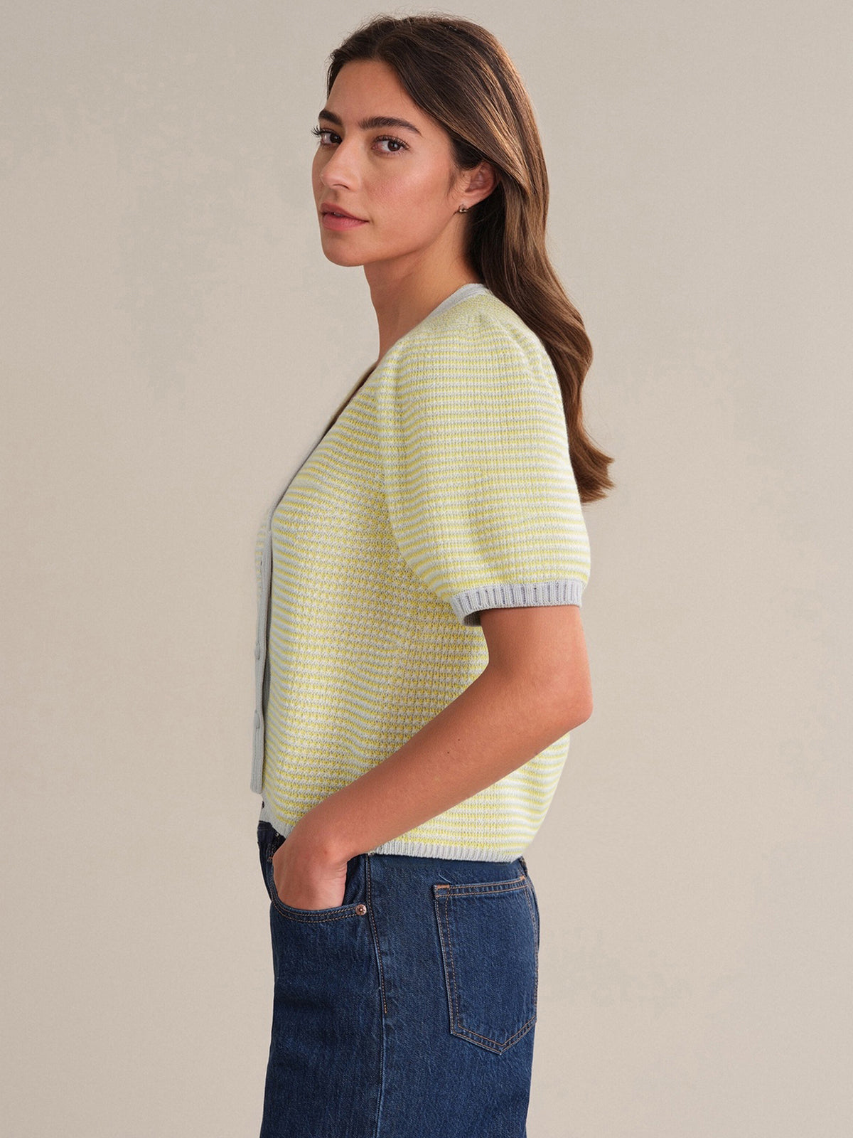 A woman with long brown hair stands in profile, wearing a light yellow 525 America V-neck short-sleeve knit cardigan with gray trim and blue jeans. Her left hand is in her pocket against a neutral background.