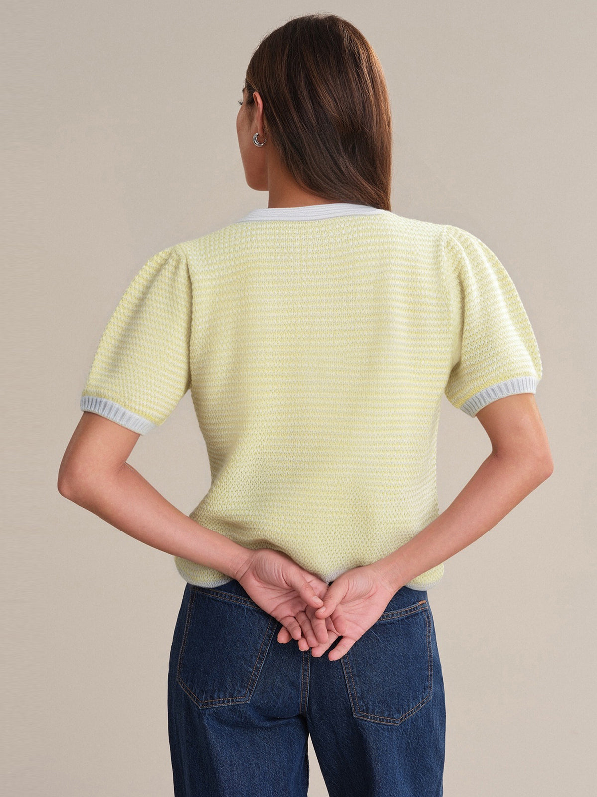 A person with long brown hair, wearing a 525 America short-sleeve yellow textured 100% cotton sweater with light gray trim and blue jeans, stands facing away with hands clasped behind their back against a plain beige background.