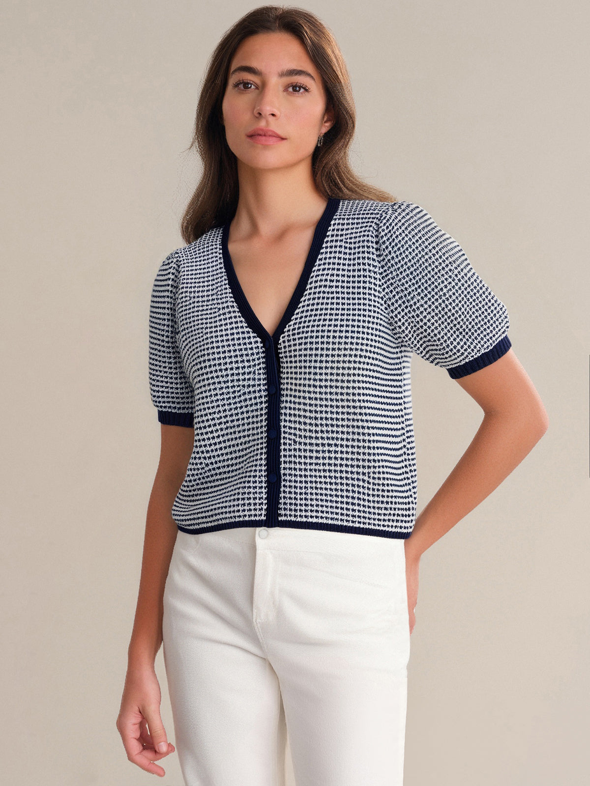 A woman with long brown hair wears a 525 America relaxed-fit, short-sleeved, V-neck navy and white checkered cardigan and high-waisted white pants, standing against a plain beige background.
