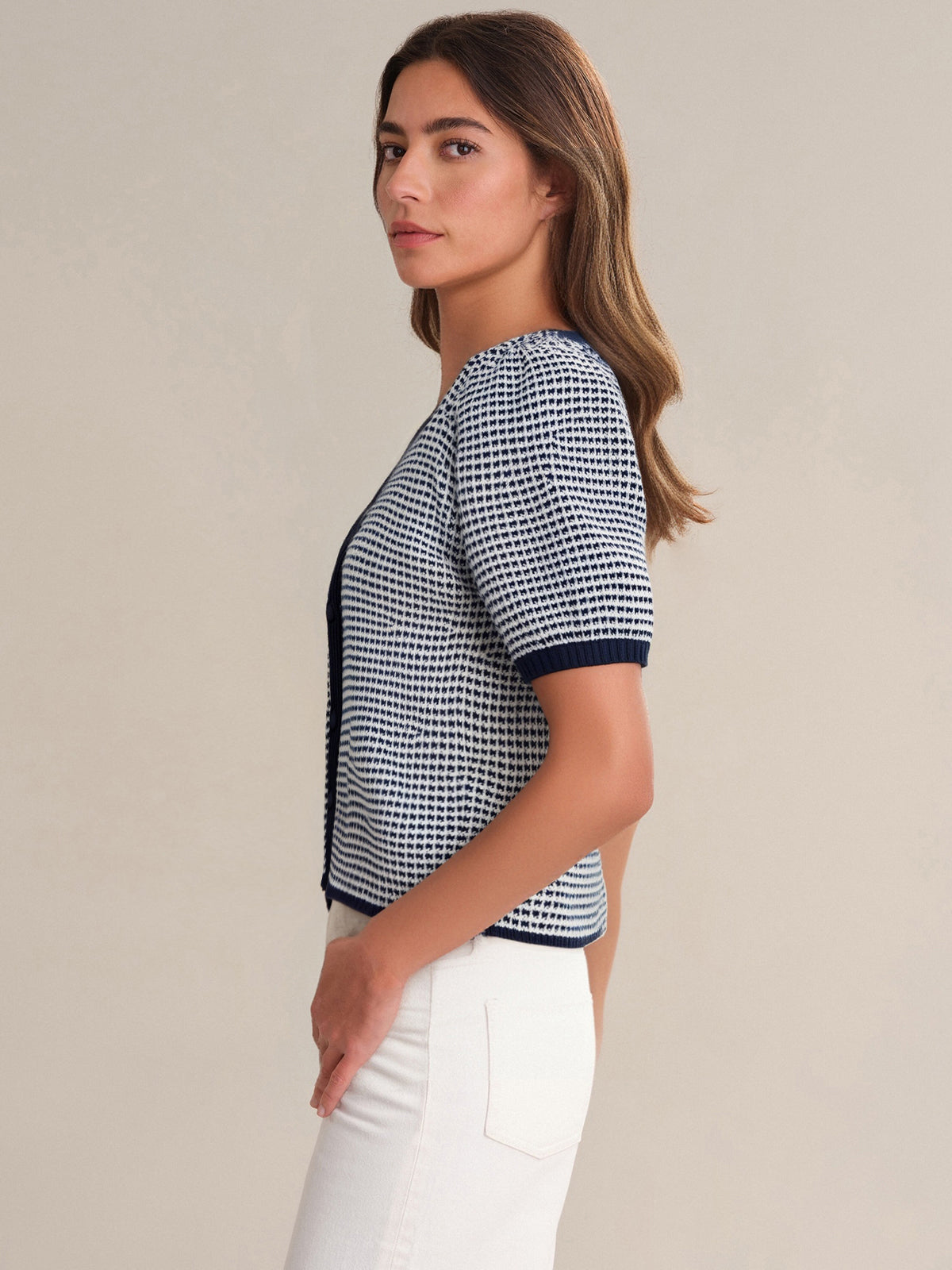 A woman with long brown hair stands in profile, wearing a 525 America V-neck navy and white checkered top with a relaxed fit and high-waisted white pants, against a plain light beige background.
