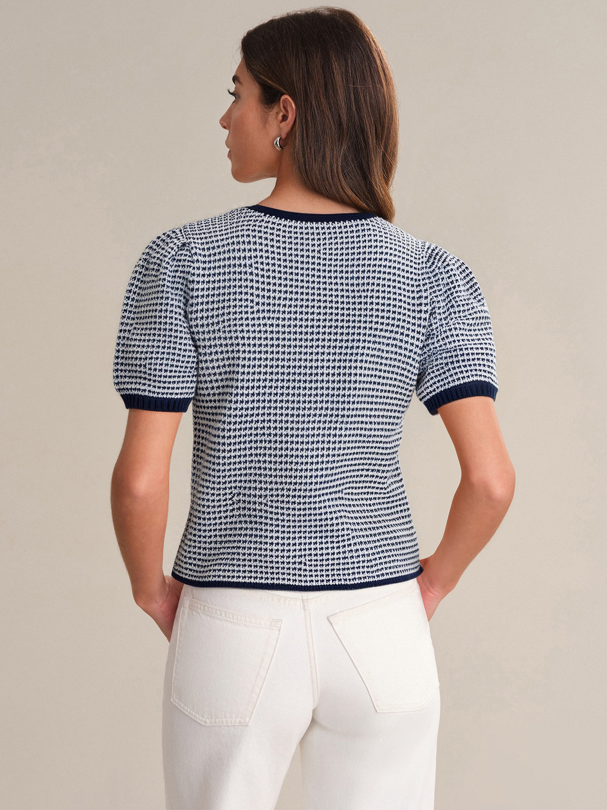 A woman stands facing away, wearing a relaxed fit, navy and white patterned knit top with navy trim by 525 America and high-waisted white pants. Her hair is down, and she stands against a plain beige background.