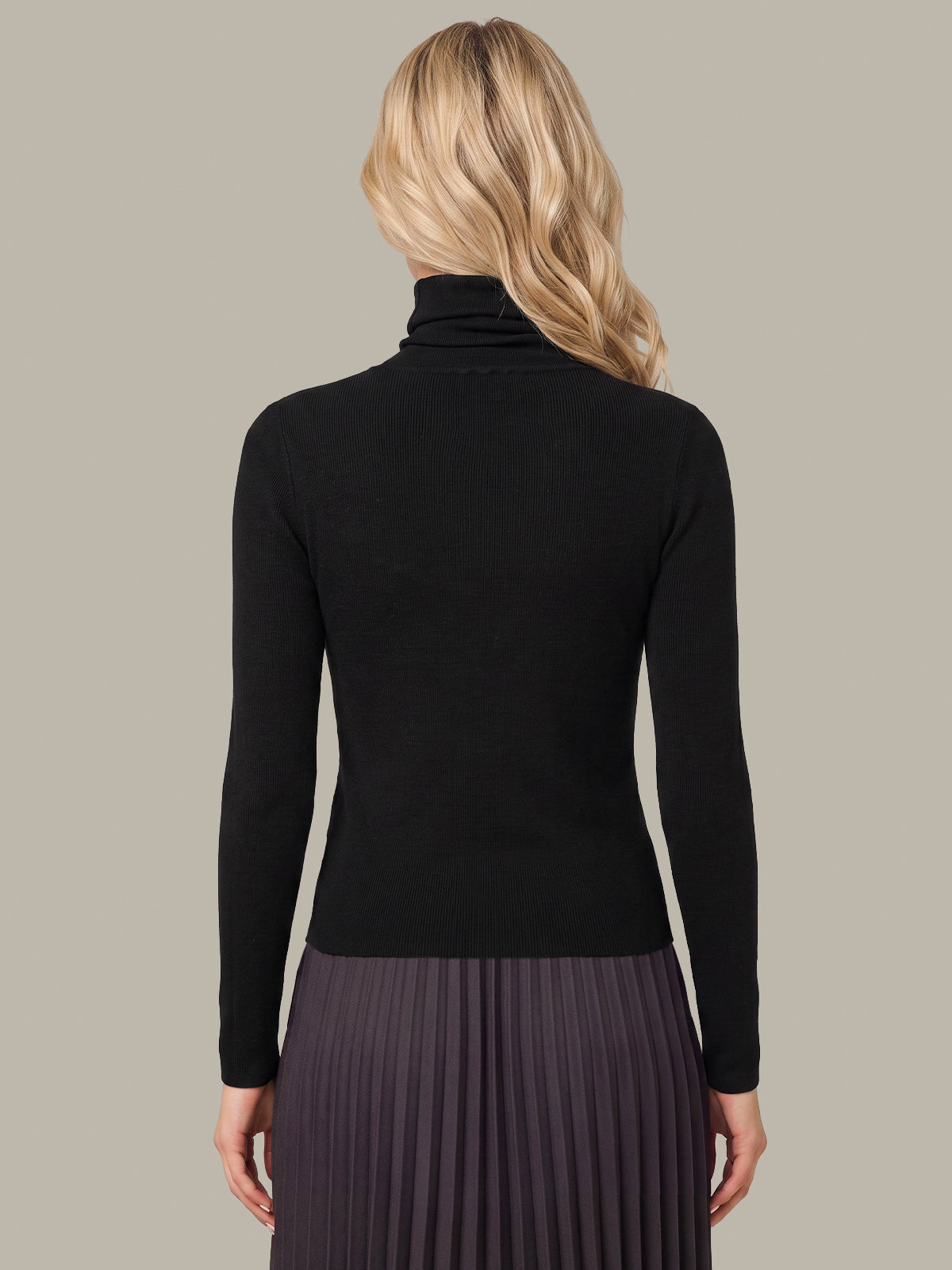 A woman shown from the back wearing a fitted black long-sleeve turtleneck top and a dark pleated skirt against a plain warm gray studio background.