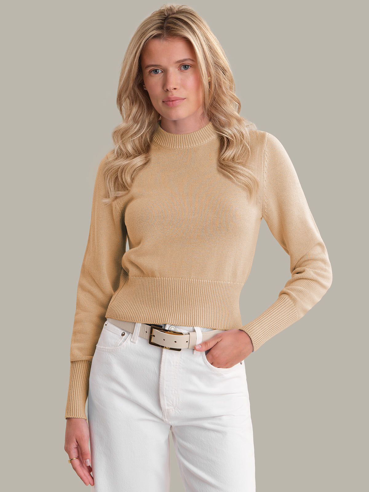Woman wearing a beige sweater and white pants against a neutral background