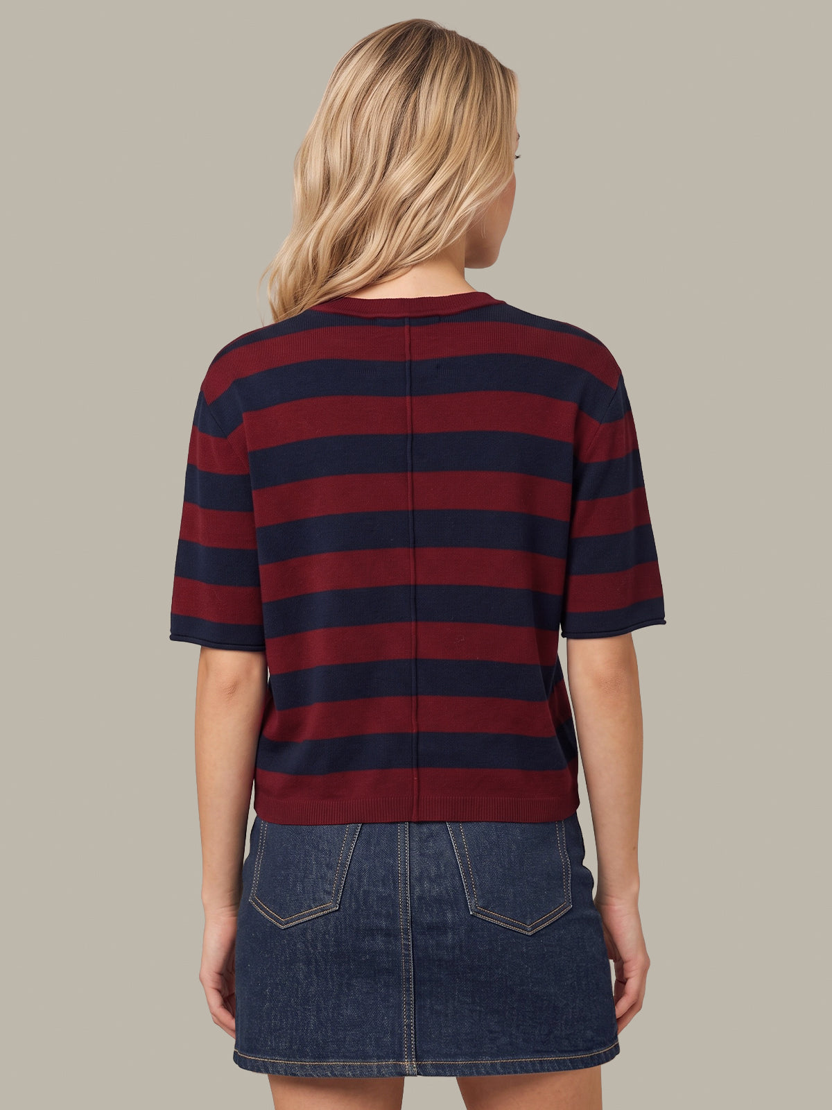 A woman shown from the back wearing a short-sleeve striped knit top with horizontal navy and burgundy red stripes, paired with a denim mini skirt, against a solid taupe background.