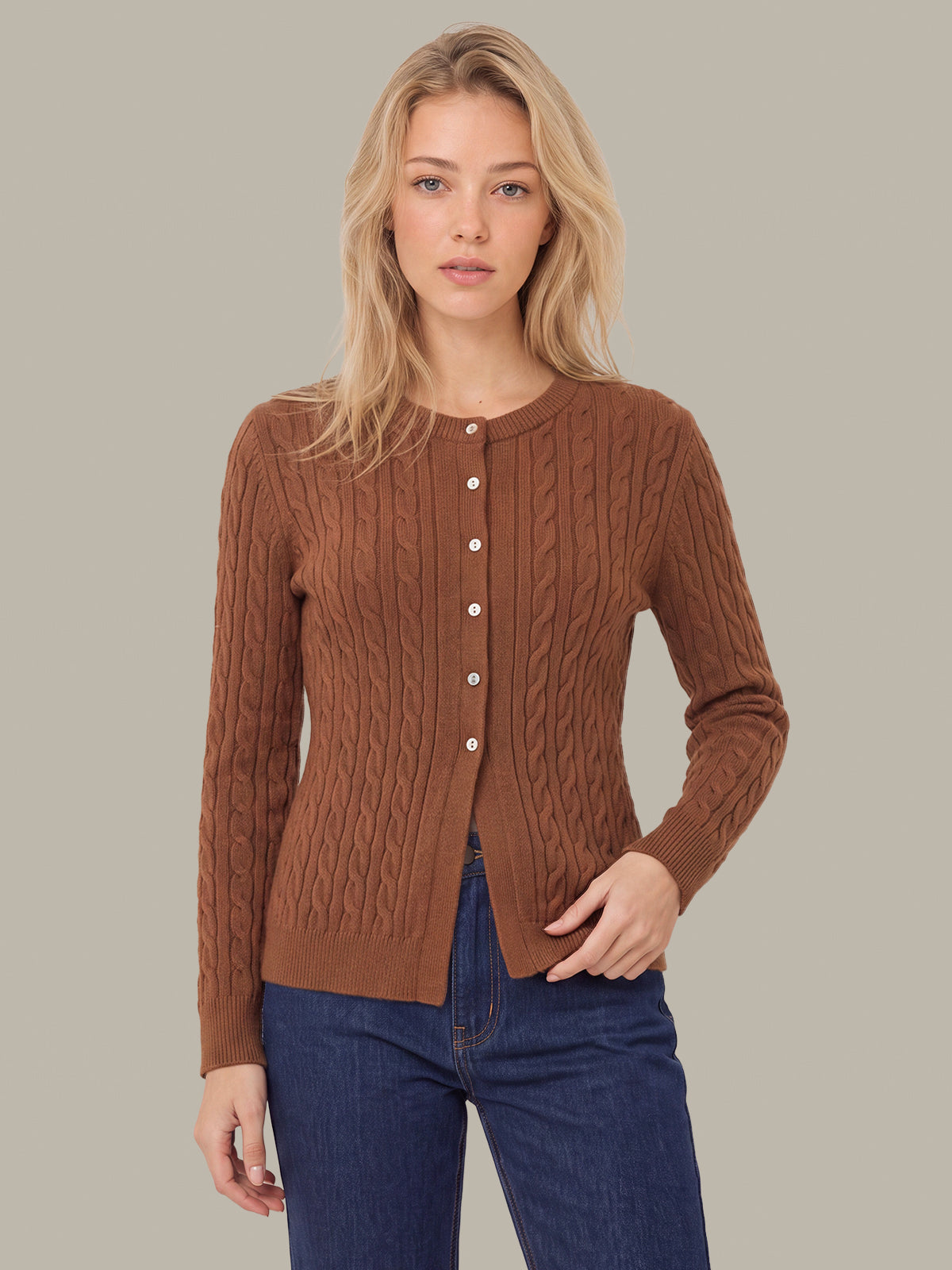 A woman modeling a brown cable-knit cardigan sweater with a button-front closure, paired with dark blue jeans, against a solid light gray background.