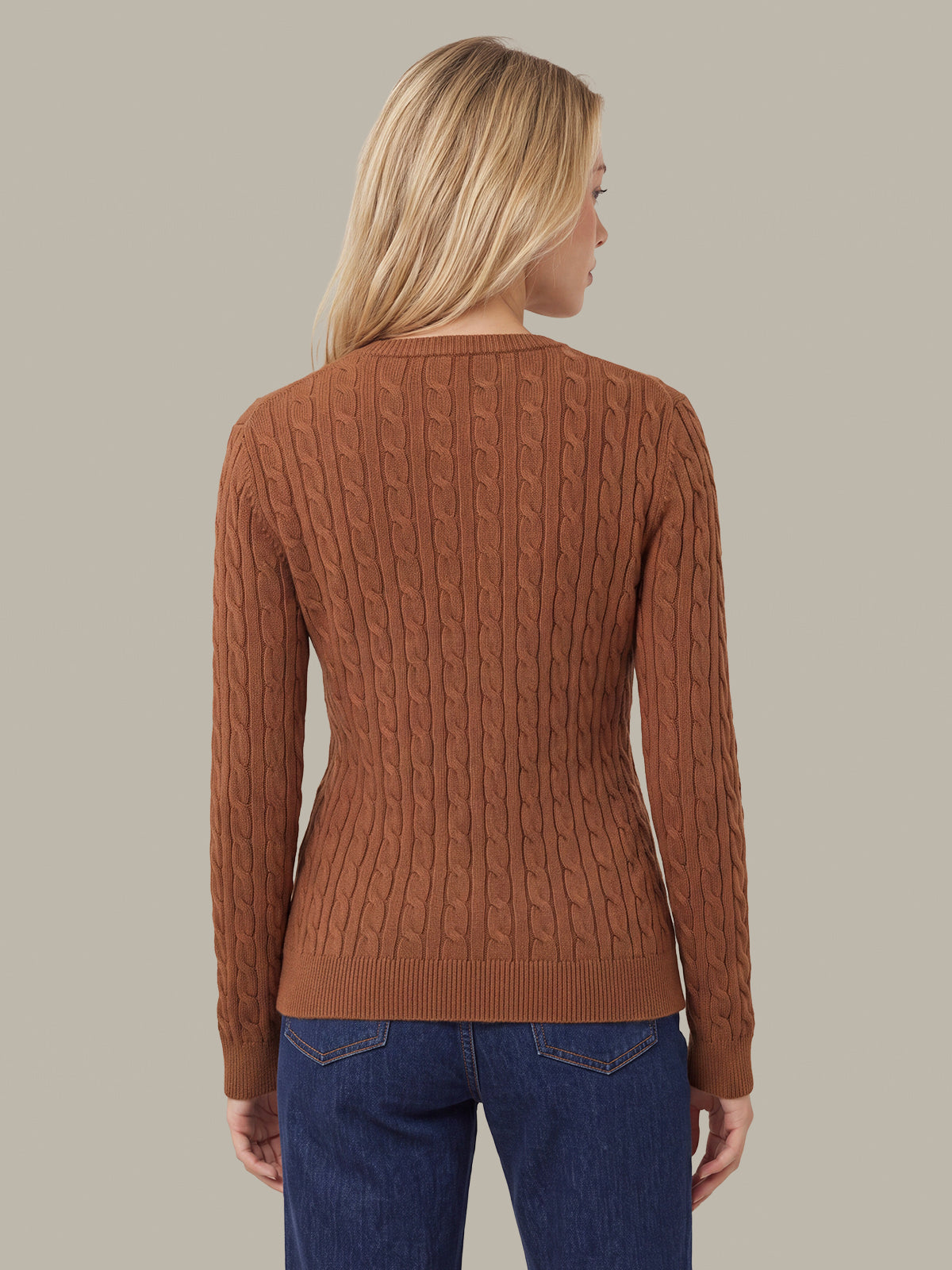 A woman shown from the back wearing a rust-brown cable-knit sweater and blue jeans against a solid light gray background.