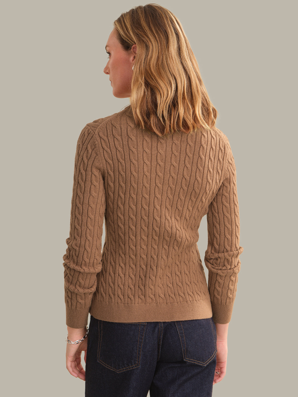 A woman shown from the back wearing a camel brown cable-knit sweater and dark blue jeans, set against a solid light gray studio background.