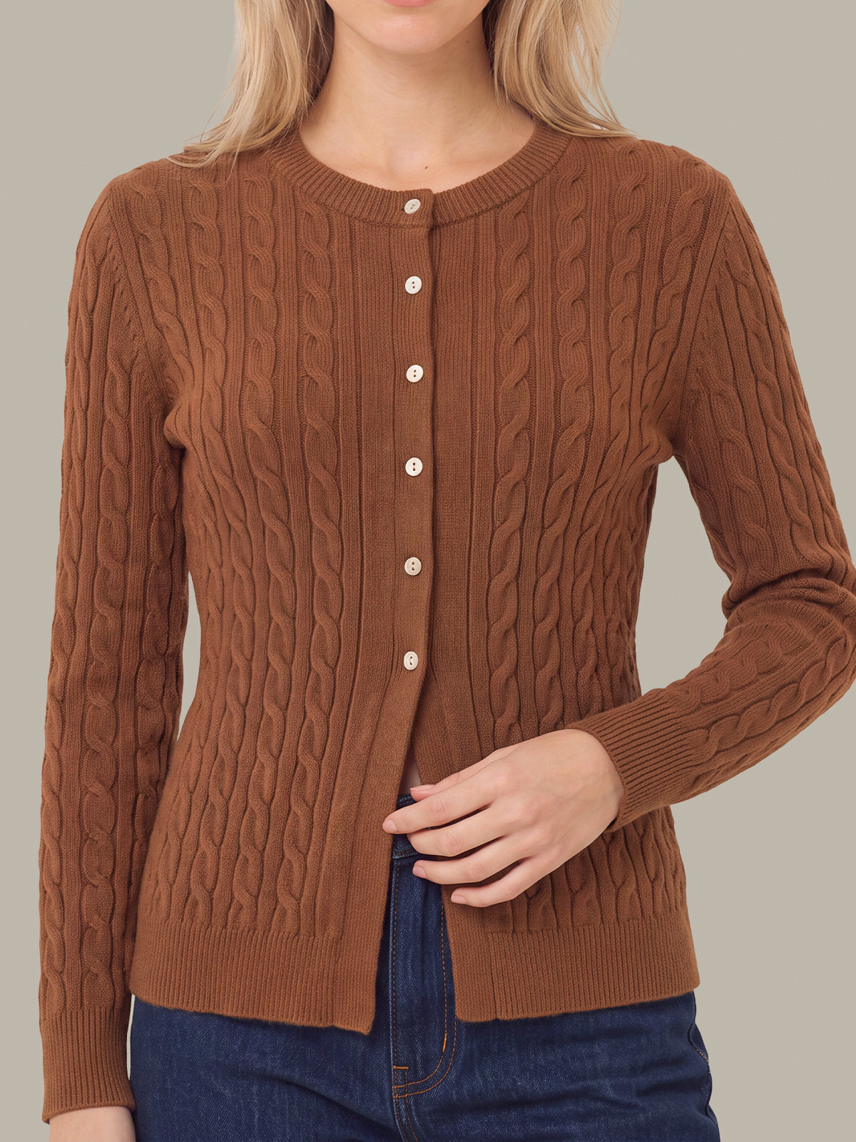 A woman wearing a brown cable-knit cardigan sweater with white buttons, paired with blue jeans, against a solid gray background.