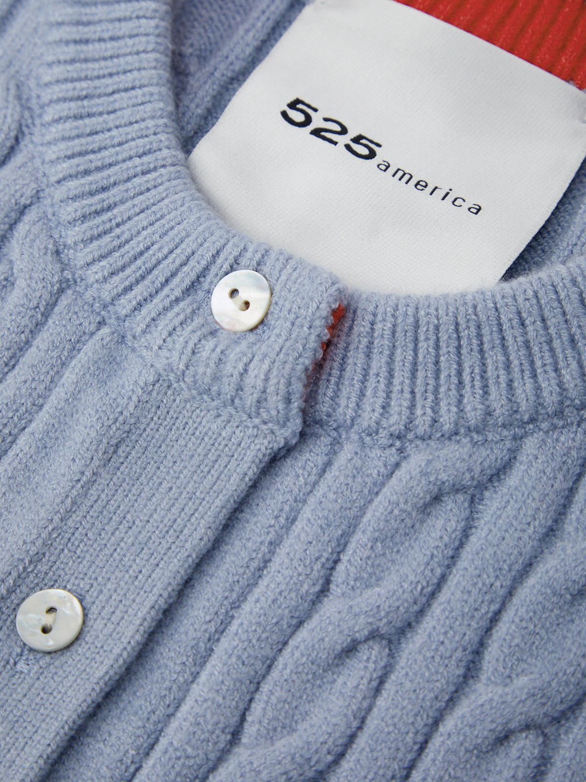 Close-up of a light blue cable-knit cardigan sweater with white buttons and a visible white neck label reading 525 america.