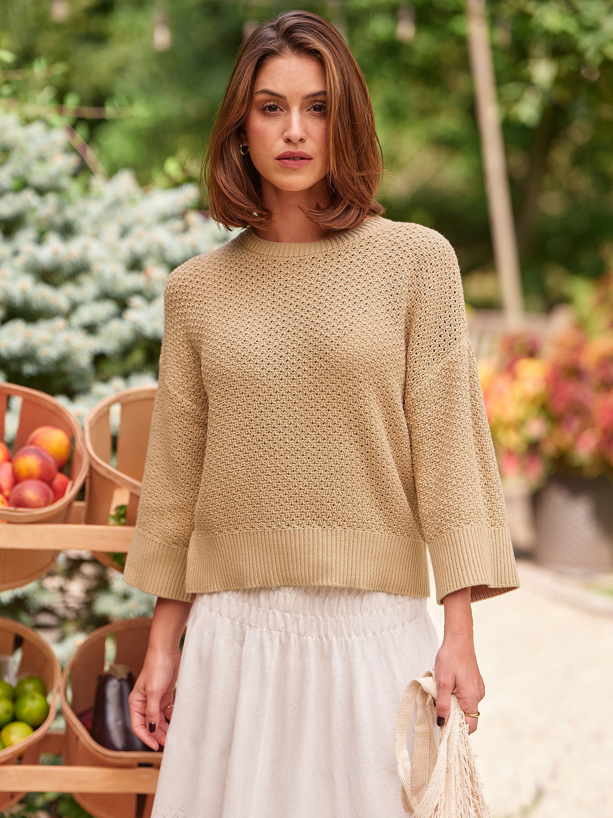 Woman wearing a beige sweater and white skirt outdoors with greenery and fruit in the background
