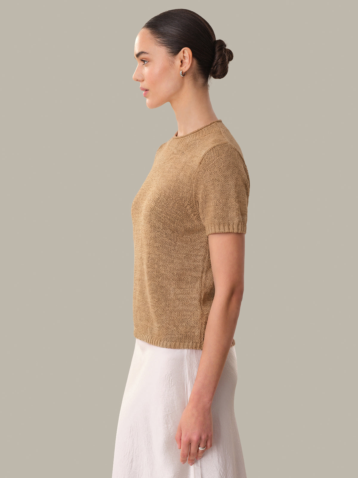 Woman wearing a beige knitted sweater and white pants against a plain background