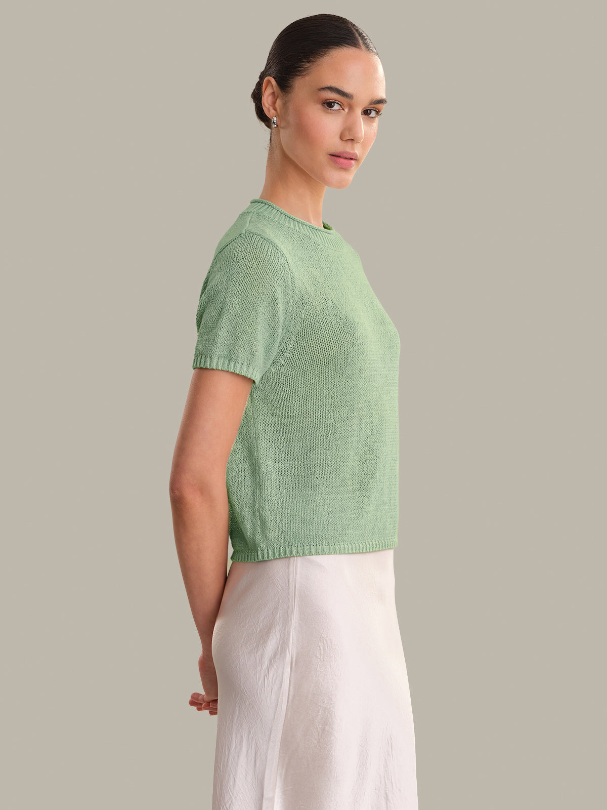 Woman wearing a green knitted top and white pants against a gray background