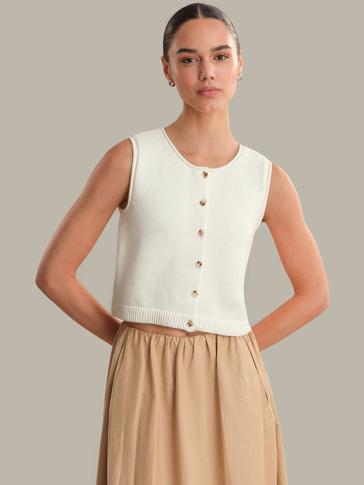Woman wearing a white sleeveless knit top and beige skirt against a neutral background