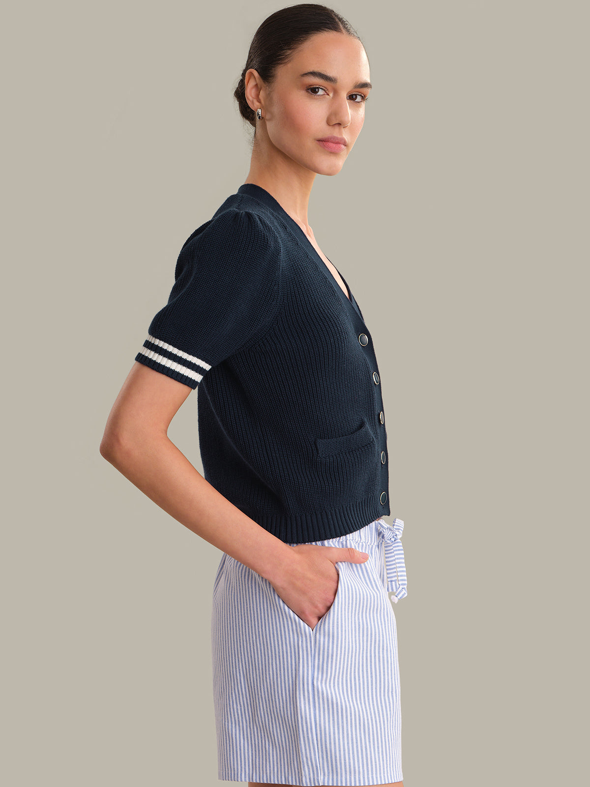 Woman wearing a navy blue cardigan with white trim and a striped skirt on a beige background