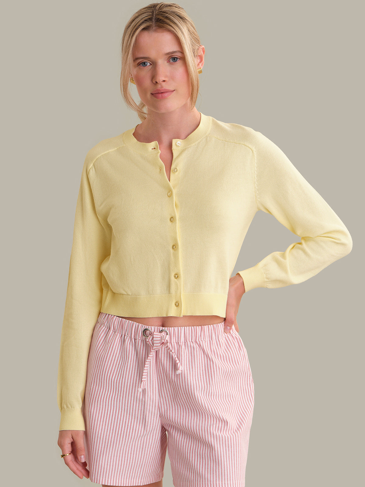 Woman wearing a light yellow cardigan over a pink top with striped pants on a neutral background