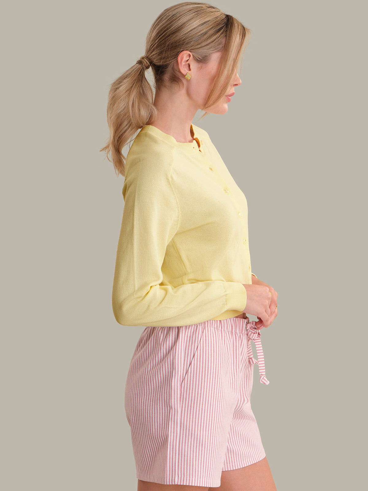 Woman wearing a yellow cardigan and pink striped shorts on a plain background