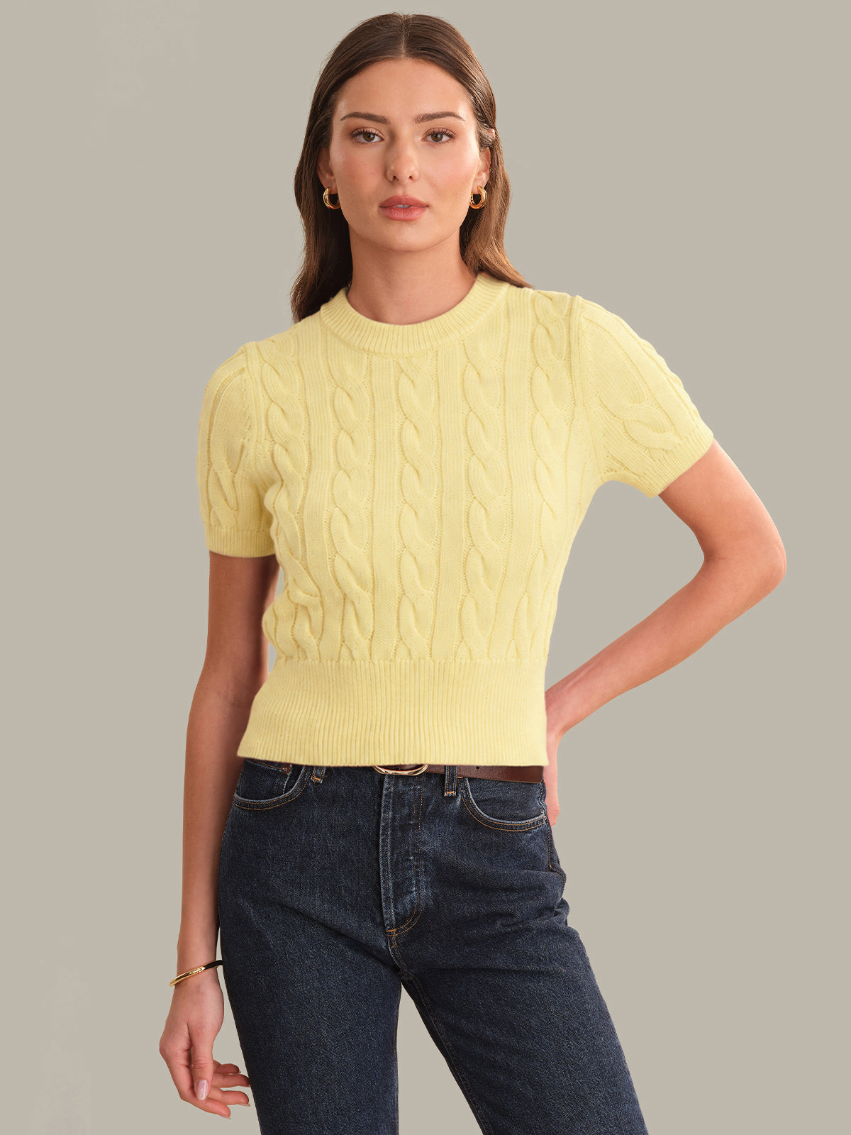 Woman wearing a yellow cable knit sweater and blue jeans on a gray background