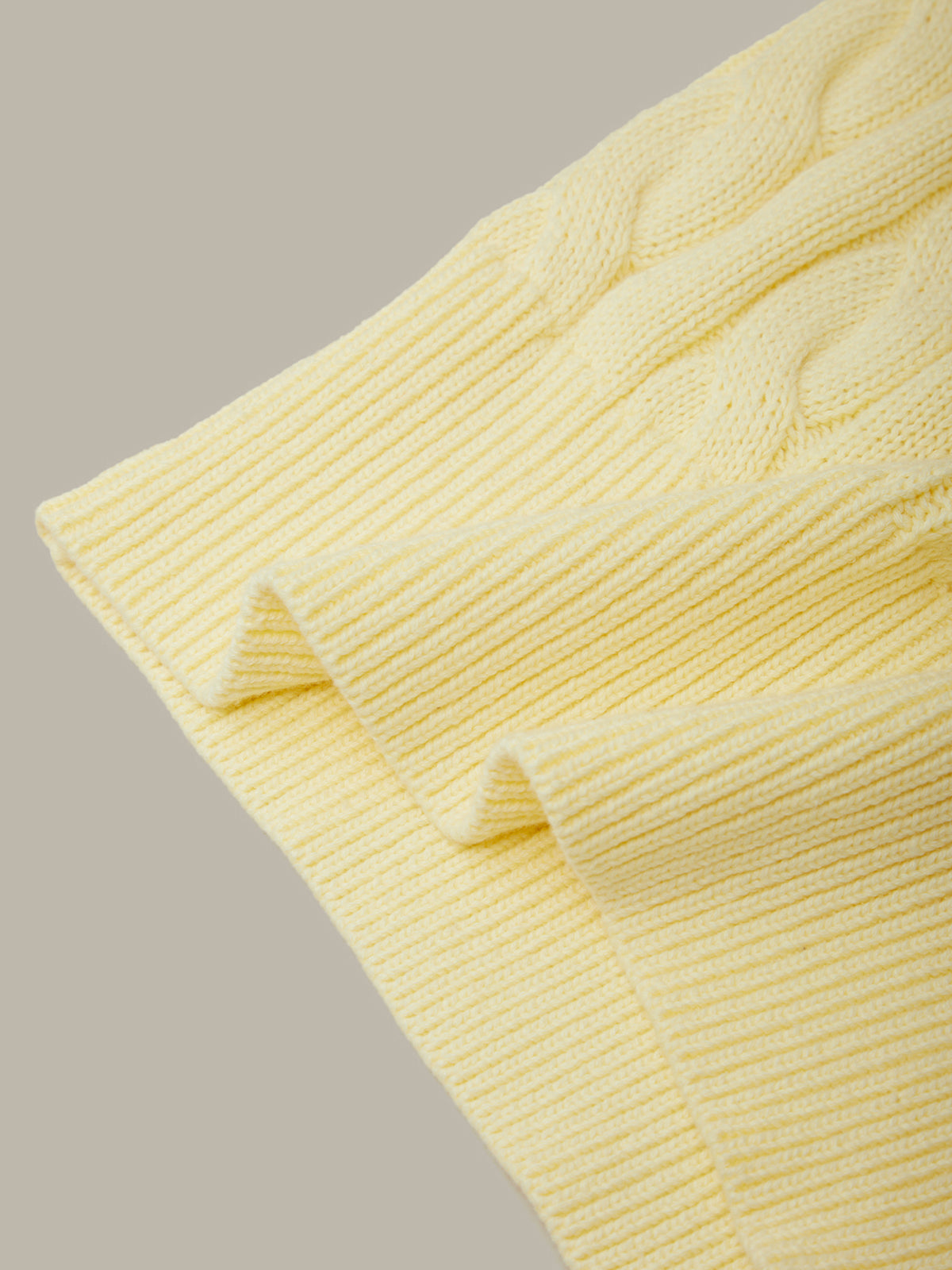 A close-up of a pale yellow cable-knit fabric with ribbed trim, folded to show texture and color against a neutral light gray background.