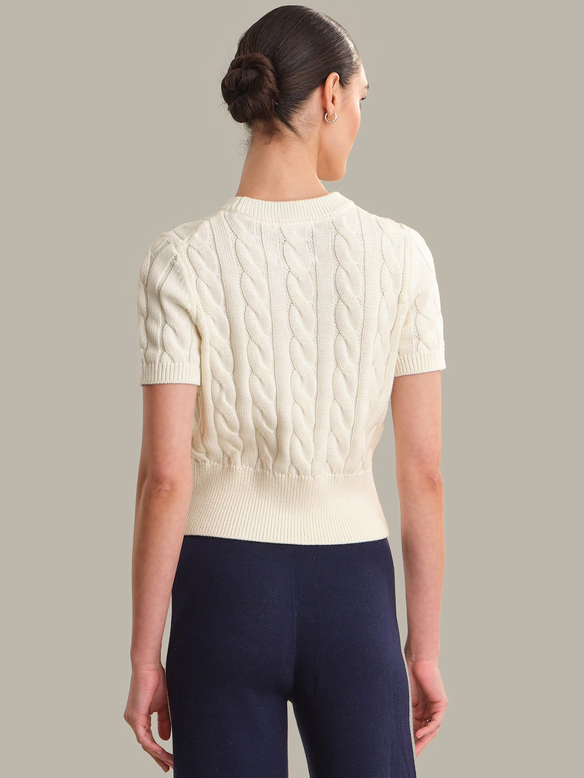 Woman wearing a cream cable knit sweater and navy pants against a gray background