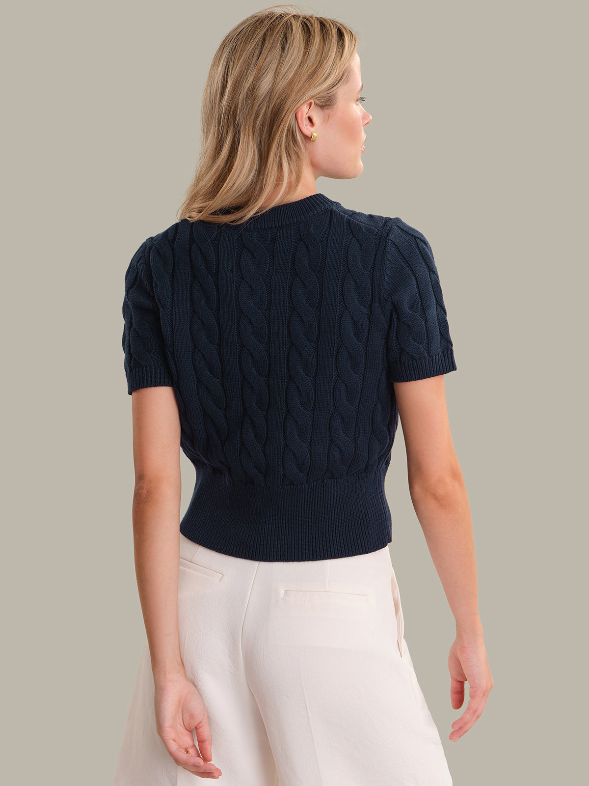 Woman wearing a navy blue cable knit top and white pants on a beige background