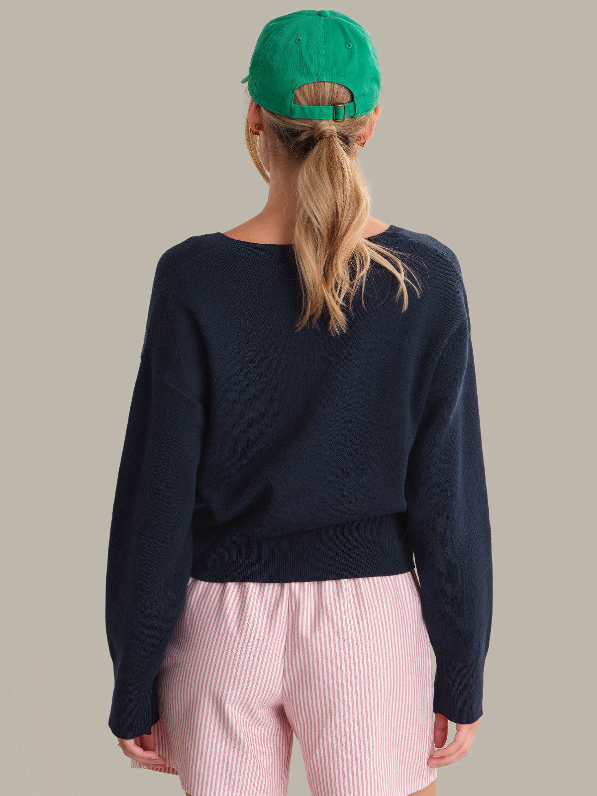 Person wearing a navy blue sweater and pink pants with a green cap on a beige background