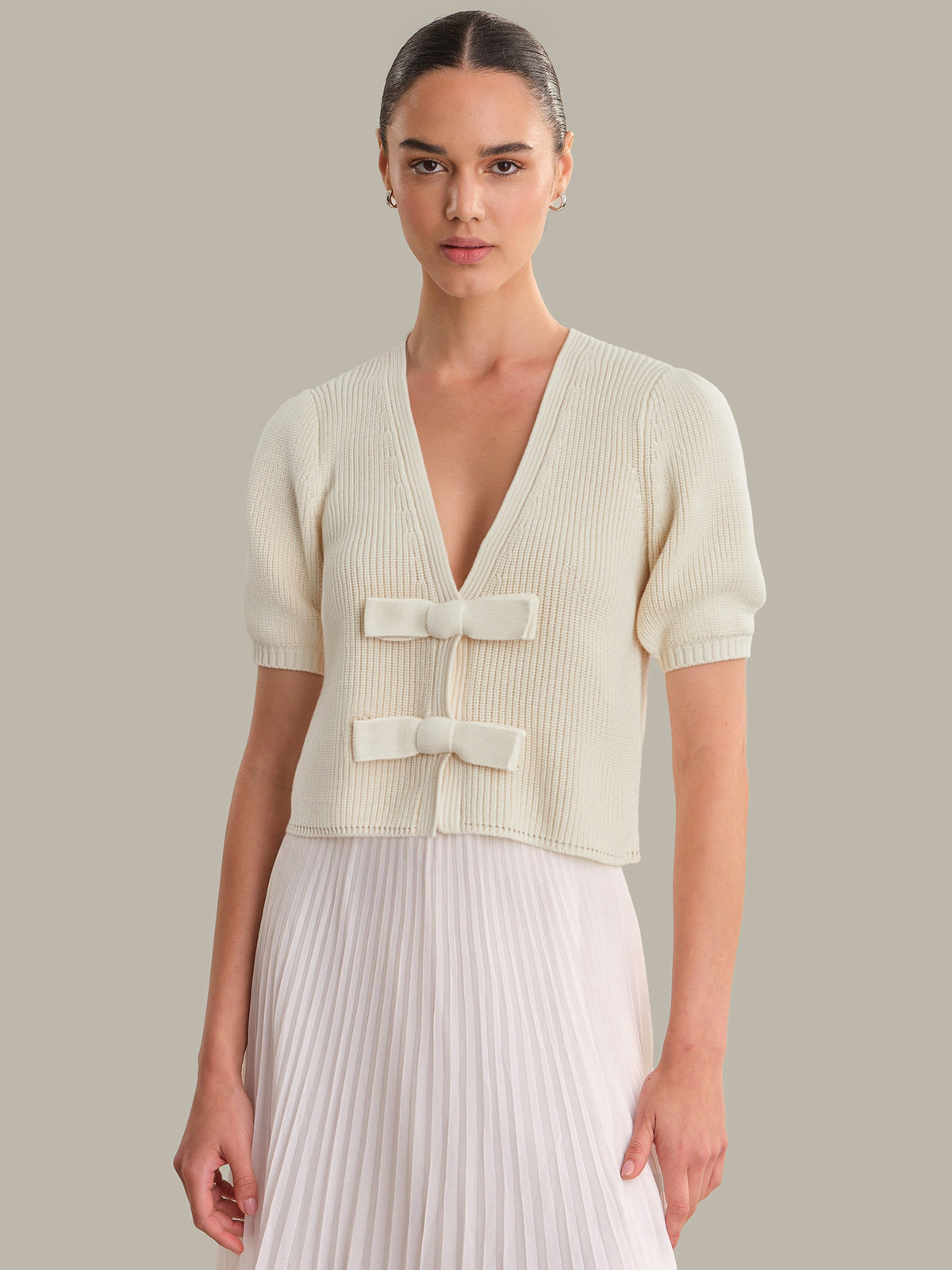 Woman wearing a cream-colored ribbed knit top with short sleeves and a pleated white skirt on a neutral background.