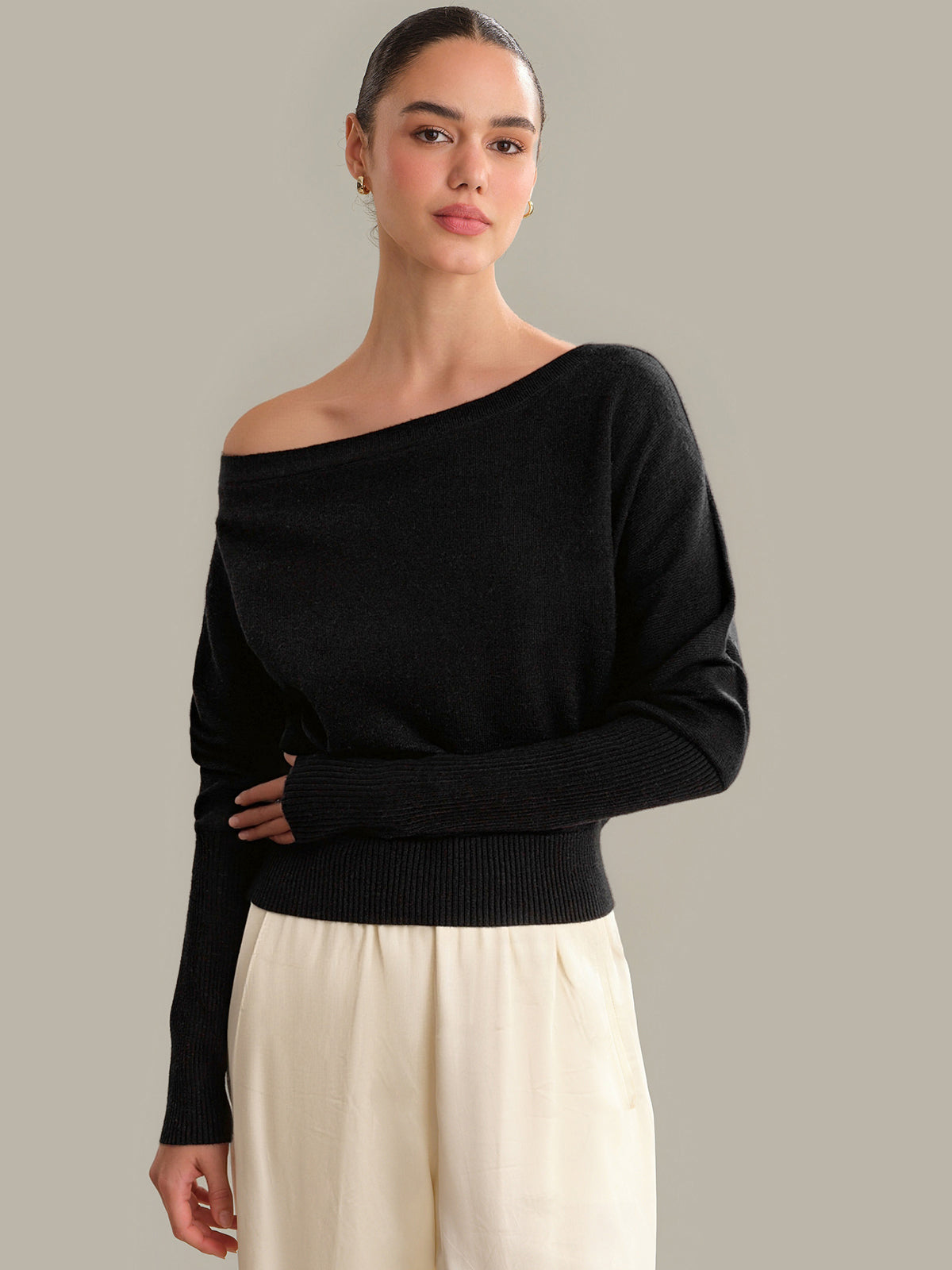 Woman wearing a black off-shoulder sweater and white pants against a gray background