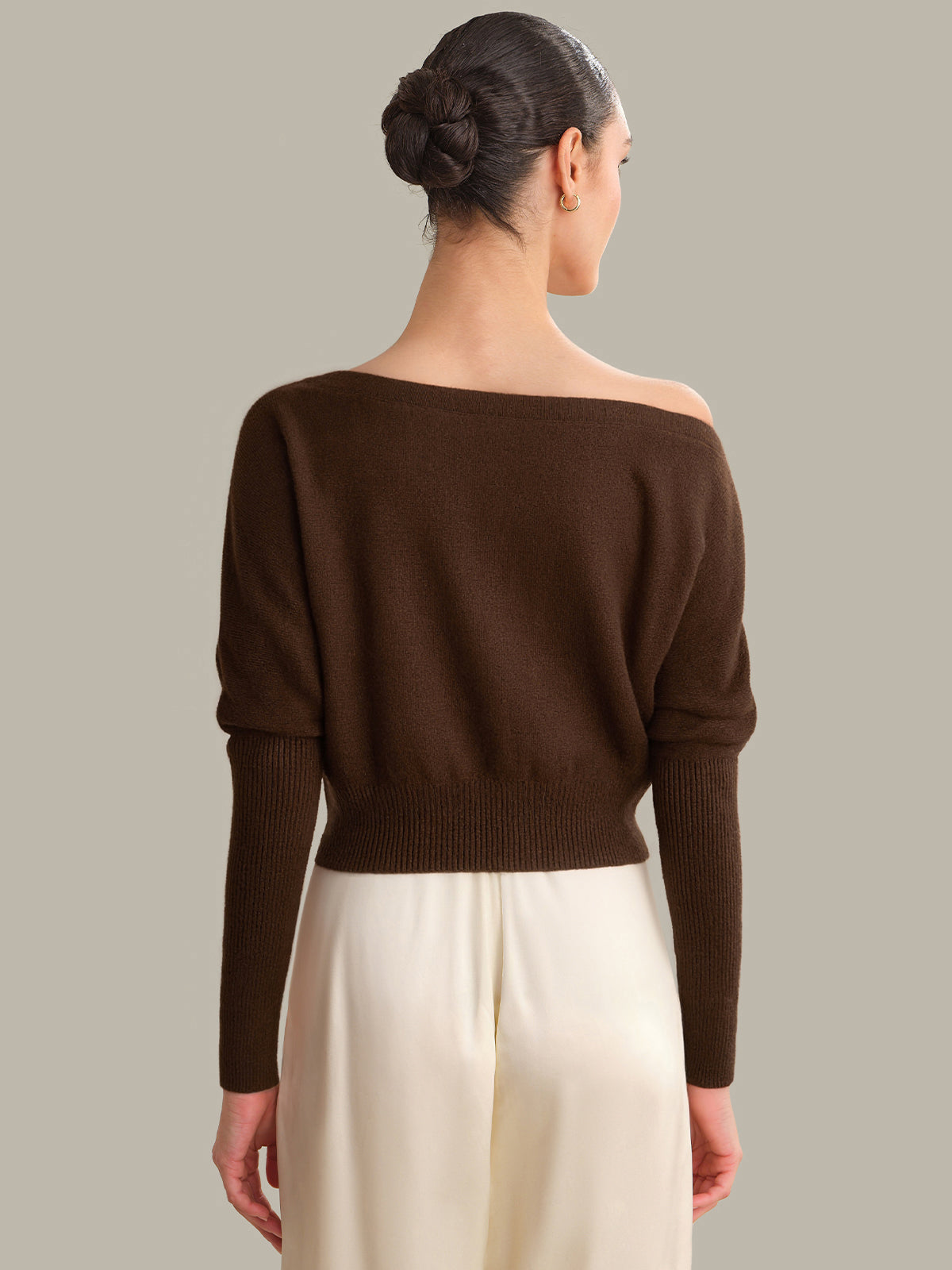 Woman wearing a brown off-shoulder sweater and white pants against a neutral background