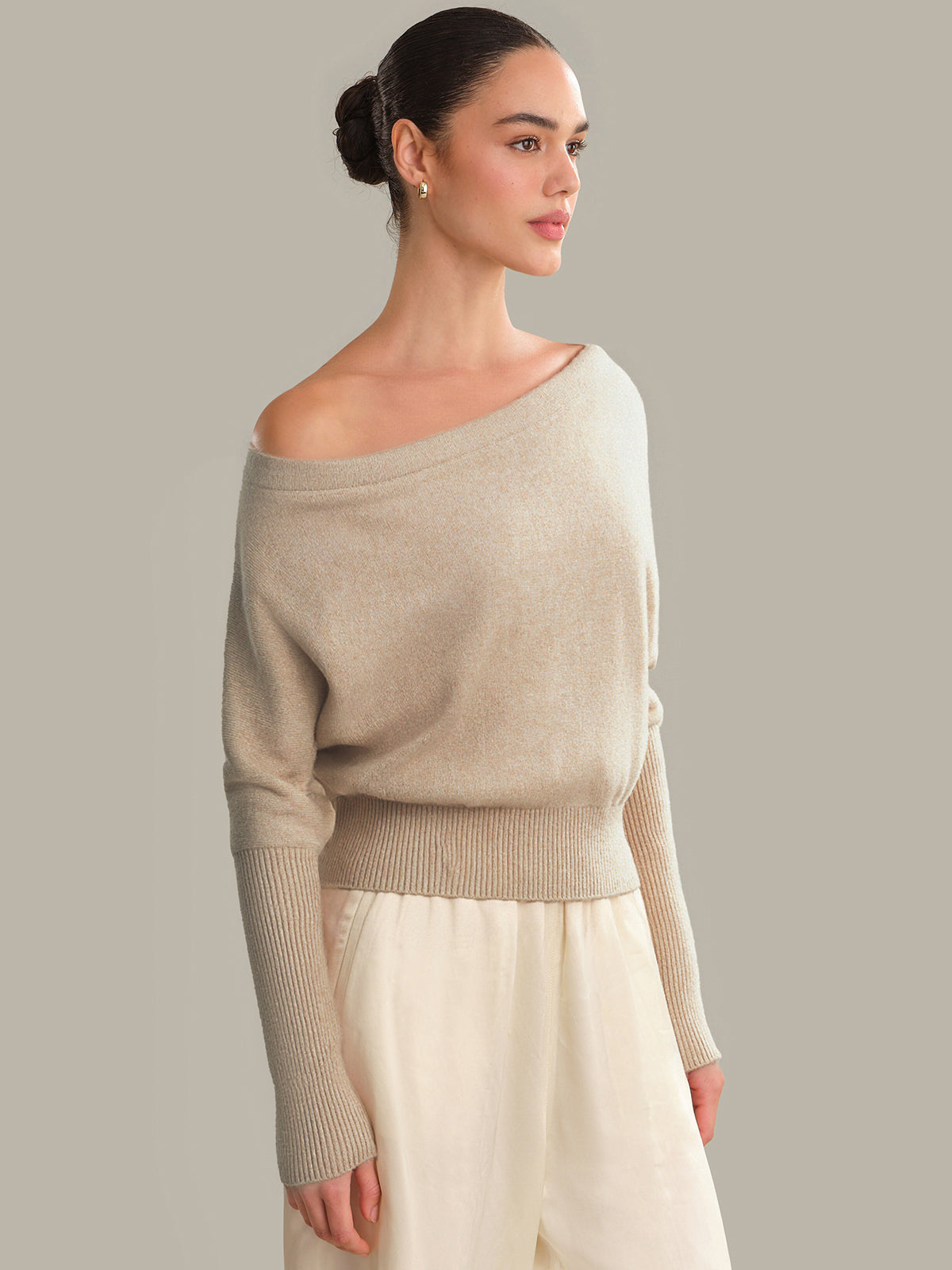 Woman wearing a beige off-shoulder sweater and white pants against a gray background