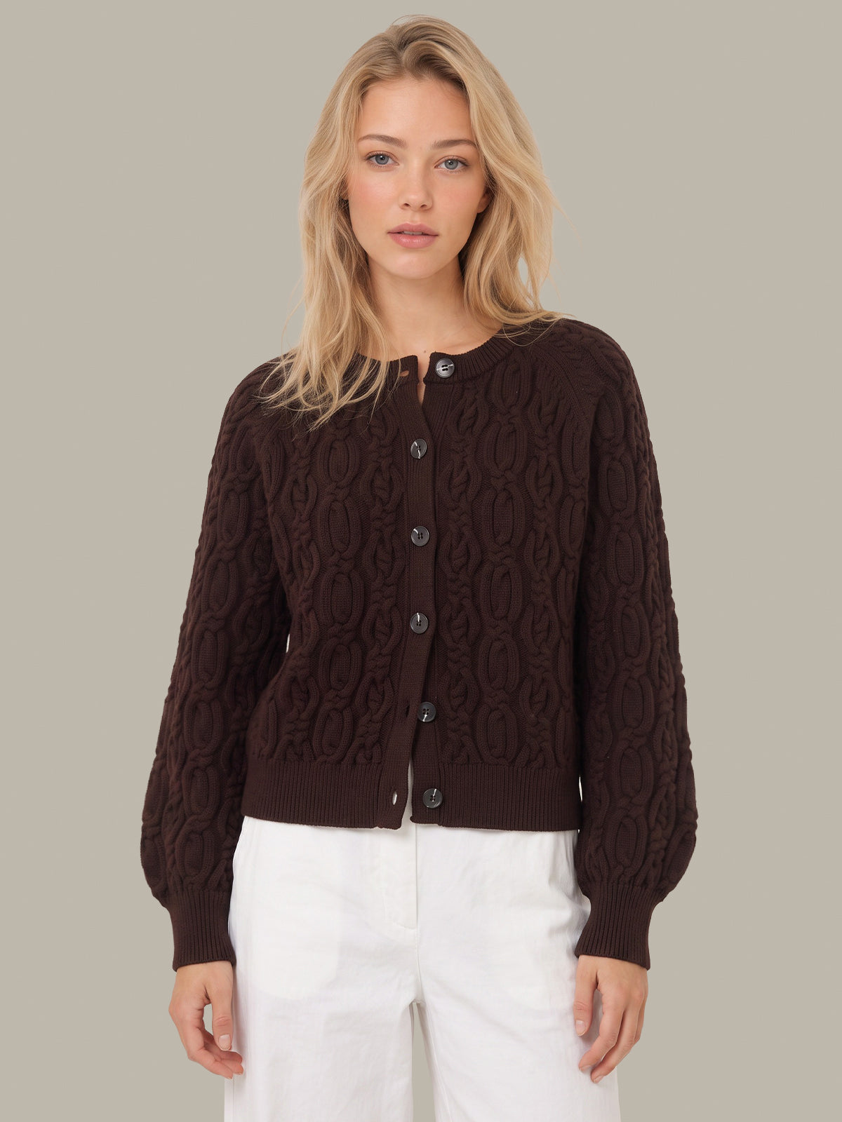 A blonde woman wearing a dark brown cable-knit cardigan with dark buttons and white pants, standing against a solid light gray background.