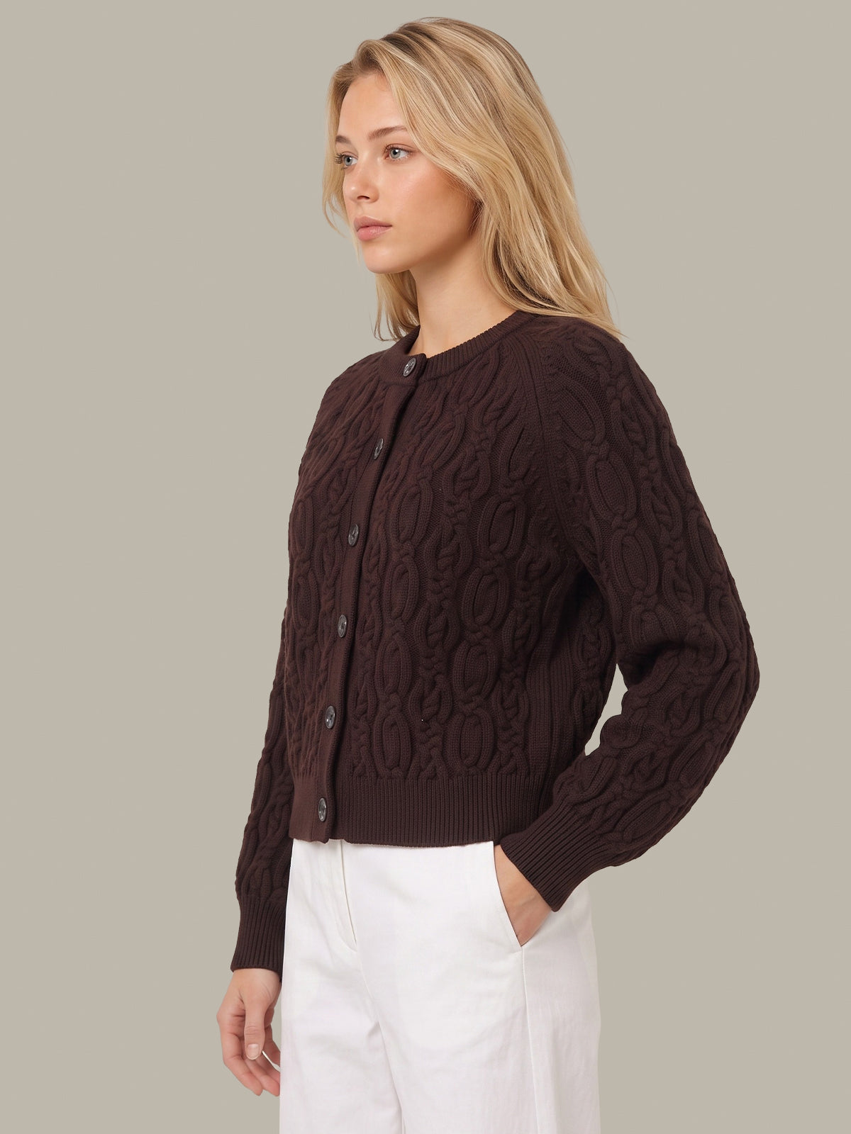 A woman wearing a dark brown cable-knit cardigan sweater and white trousers, shown from a side profile against a solid light gray background.